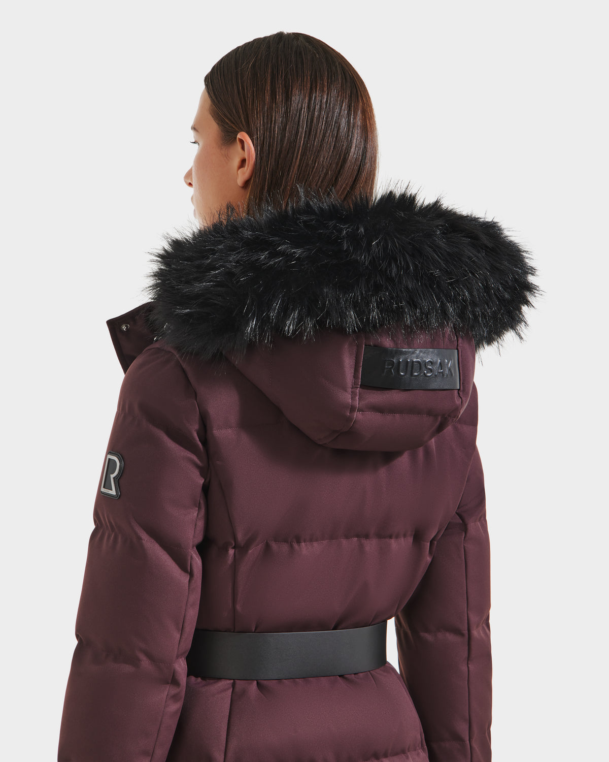 A person with straight brown hair is seen from the back wearing the Rudsak IZABELLA H - 8125644 Bordeaux women’s cold-weather puffer coat with RDS-certified down, black faux fur hood, R patch on the sleeve, and a wide black waist belt.