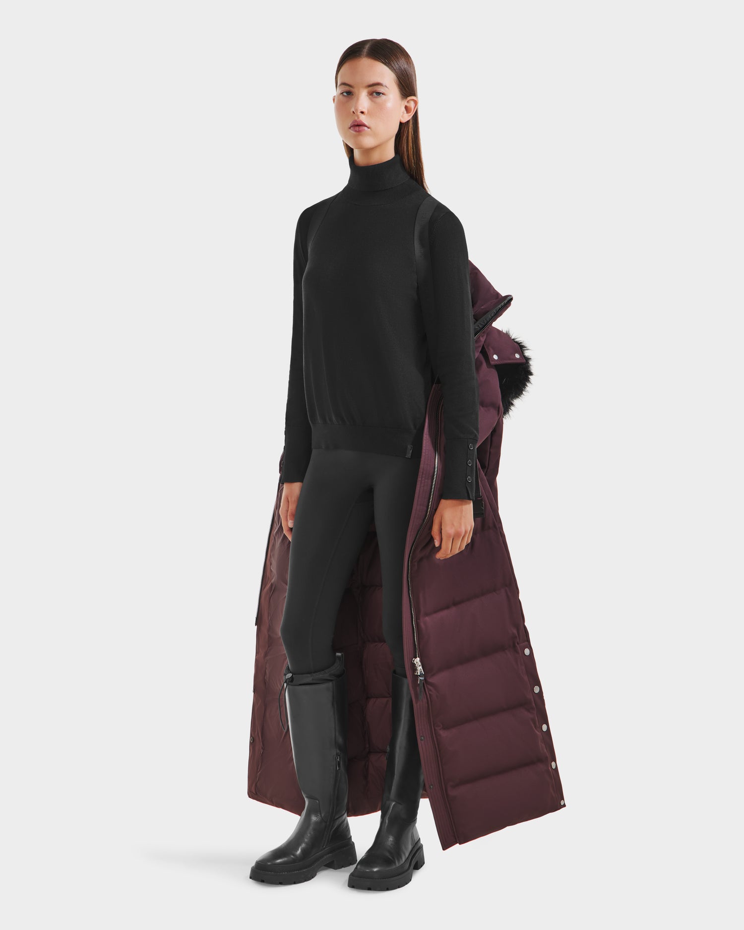 A woman stands against a plain white background wearing the Rudsak IZABELLA H - 8125644 Bordeaux, a long maroon coat with RDS-certified down, draped elegantly over her shoulders atop a black turtleneck, leggings, and knee-high boots.