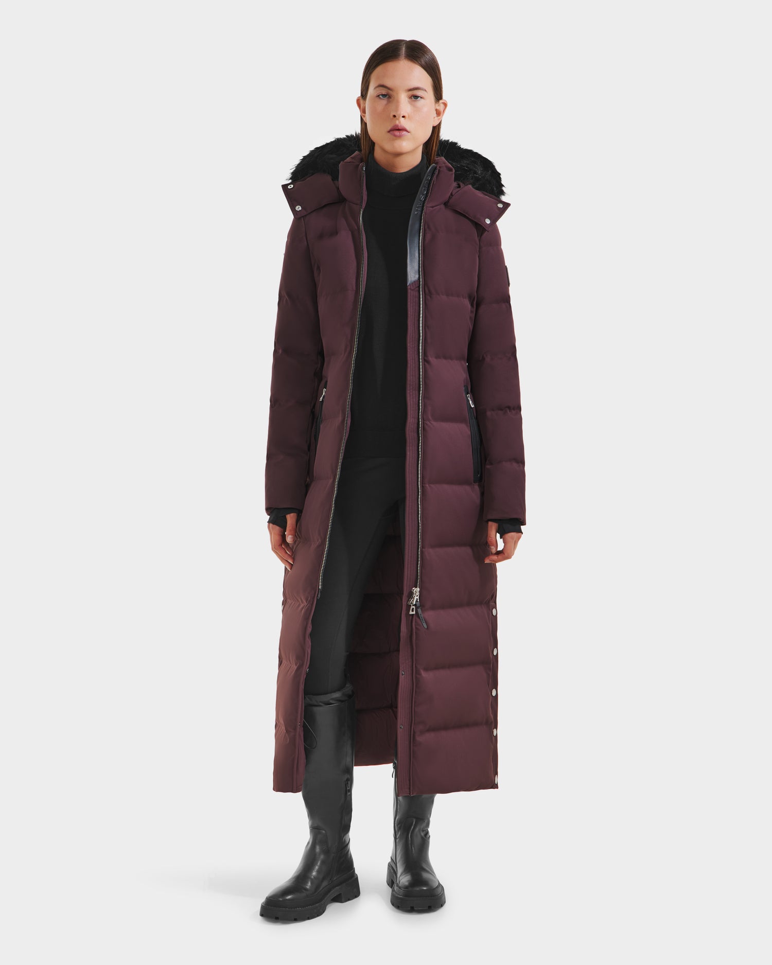 A woman faces forward, wearing the Rudsak IZABELLA H - 8125644 Bordeaux long maroon puffer with a hood, RDS-certified down filling, black pants, turtleneck, and knee-high boots against a white background.