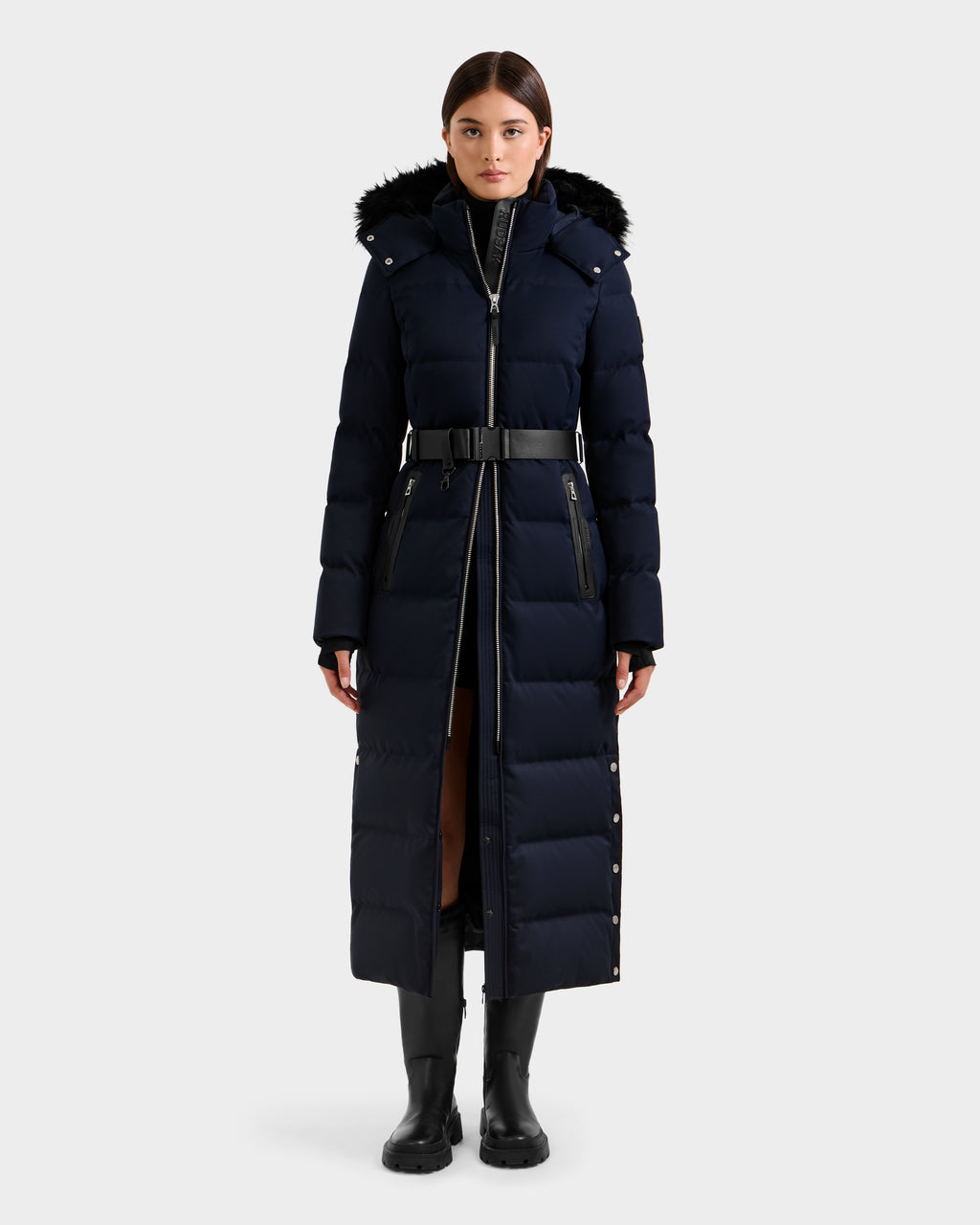 A woman faces forward wearing the Rudsak IZABELLA H - 8125644 Navy puffer with a black belt and fur-trimmed hood, styled with black boots and gloves, set against a plain light gray background.