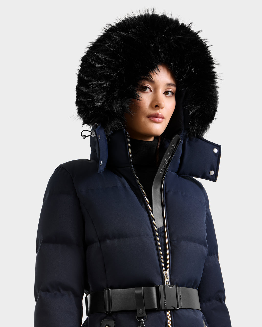 A woman wears the Rudsak IZABELLA H - 8125644 Navy puffer coat, featuring a black faux fur-lined hood, black belt, and silver zipper, set against a plain light gray background.