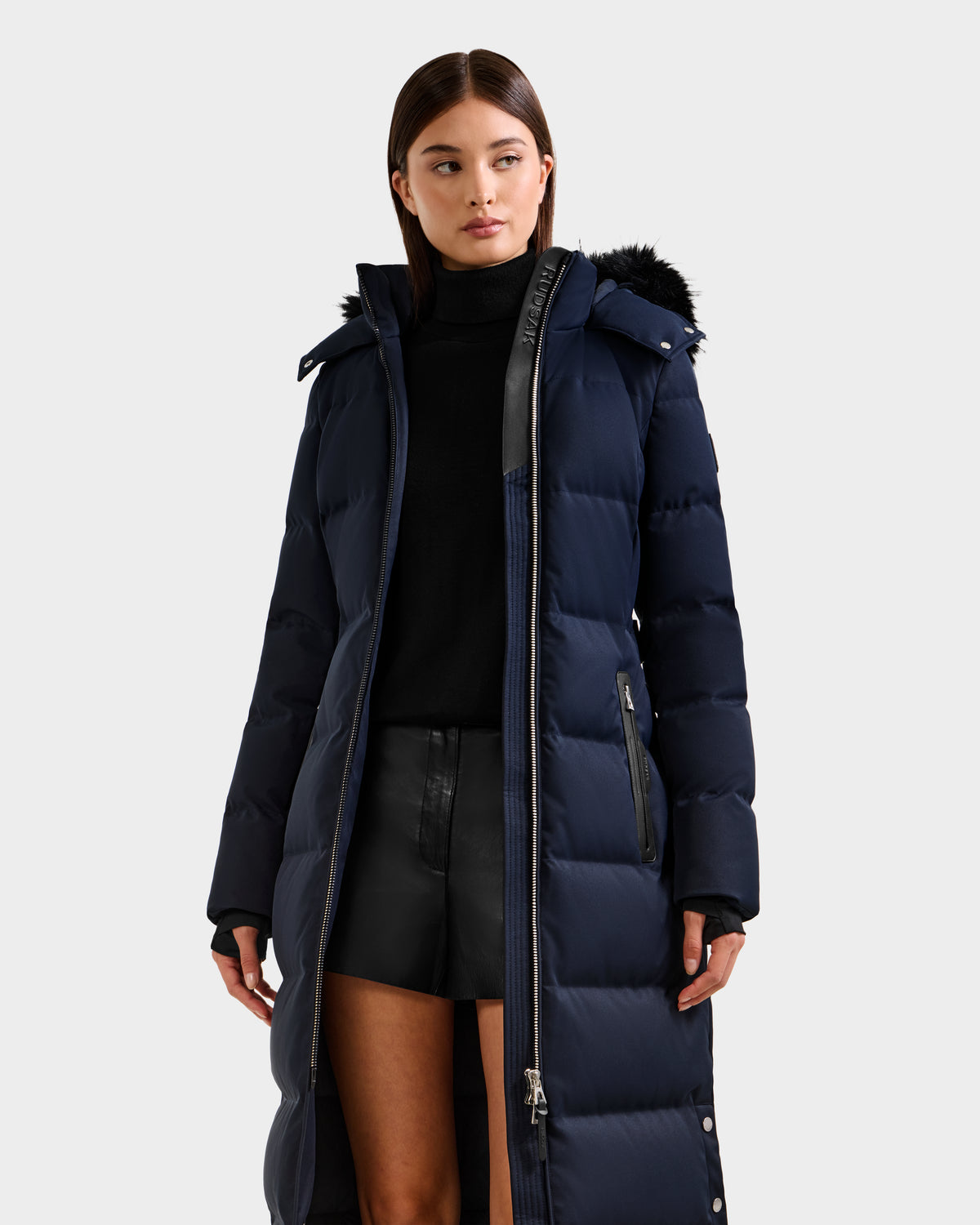 A woman wears the Rudsak IZABELLA H - 8125644 Navy puffer with a black faux fur-trimmed hood, paired with a black turtleneck and leather shorts, standing against a plain light grey background.
