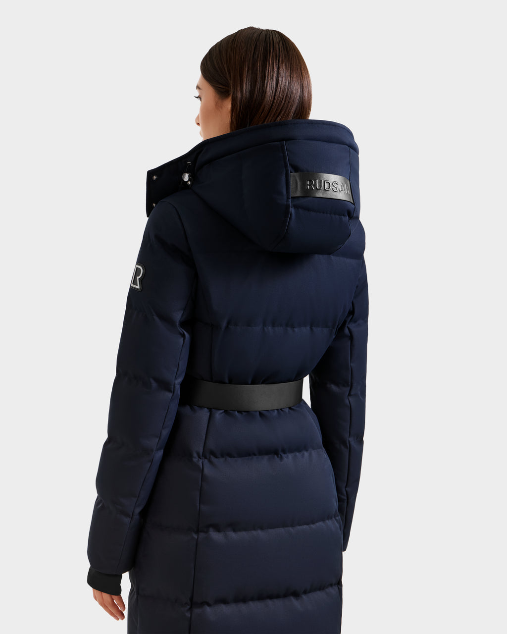 A woman with straight brown hair, seen from behind, wears the Rudsak IZABELLA H - 8125644 Navy women's puffer winter jacket with a hood, RUDSAK patch on the hood, black waist belt, and a light gray background.