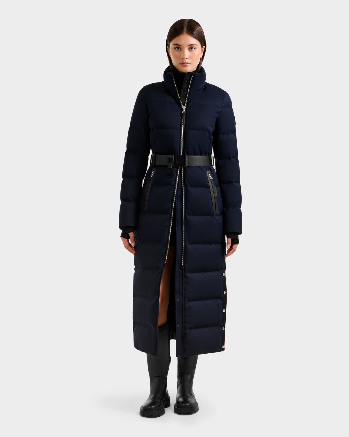 A woman faces forward wearing the Rudsak IZABELLA H - 8125644 Navy, a long women’s puffer with a belted waist, high collar, visible zippers, snap buttons along the front and side, paired with black boots.