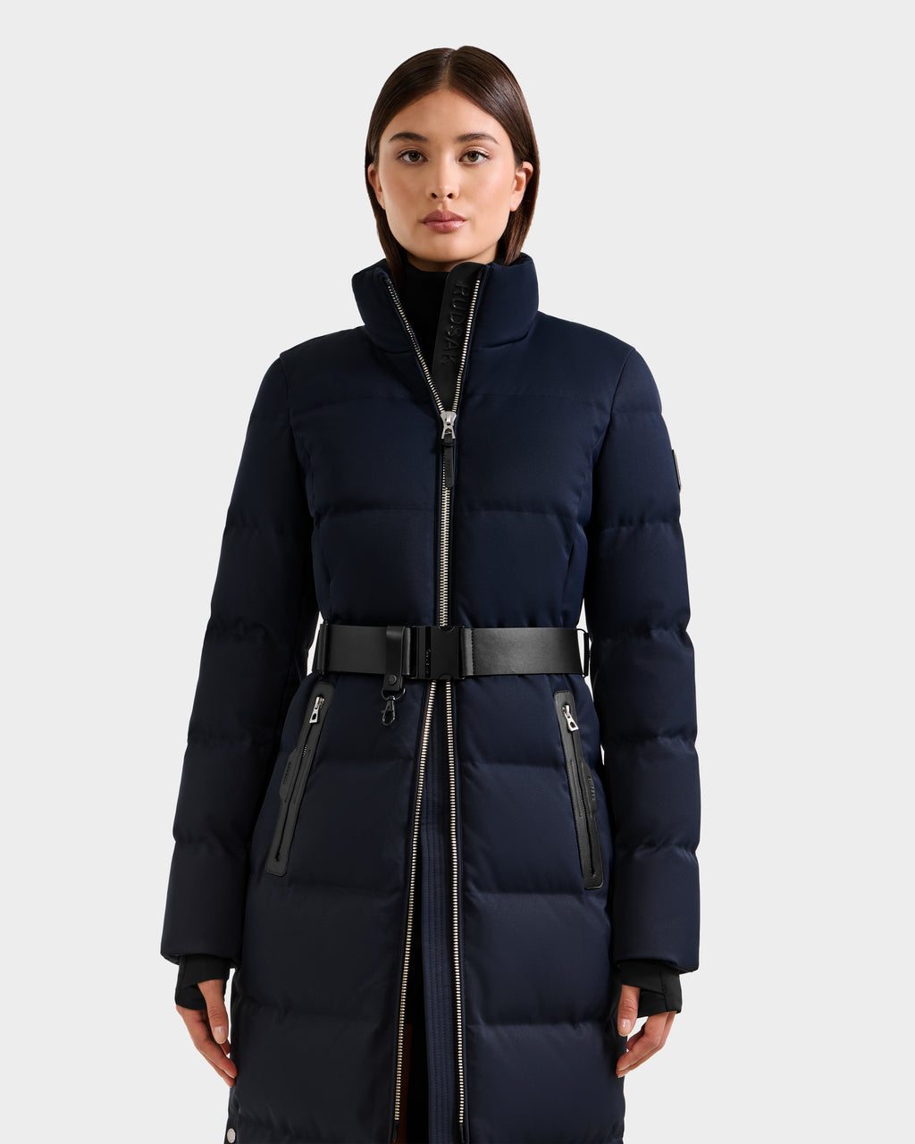 A woman faces forward against a plain light gray background, wearing the Rudsak IZABELLA H - 8125644 Navy puffer coat featuring a high collar, black belt, and zippered pockets.