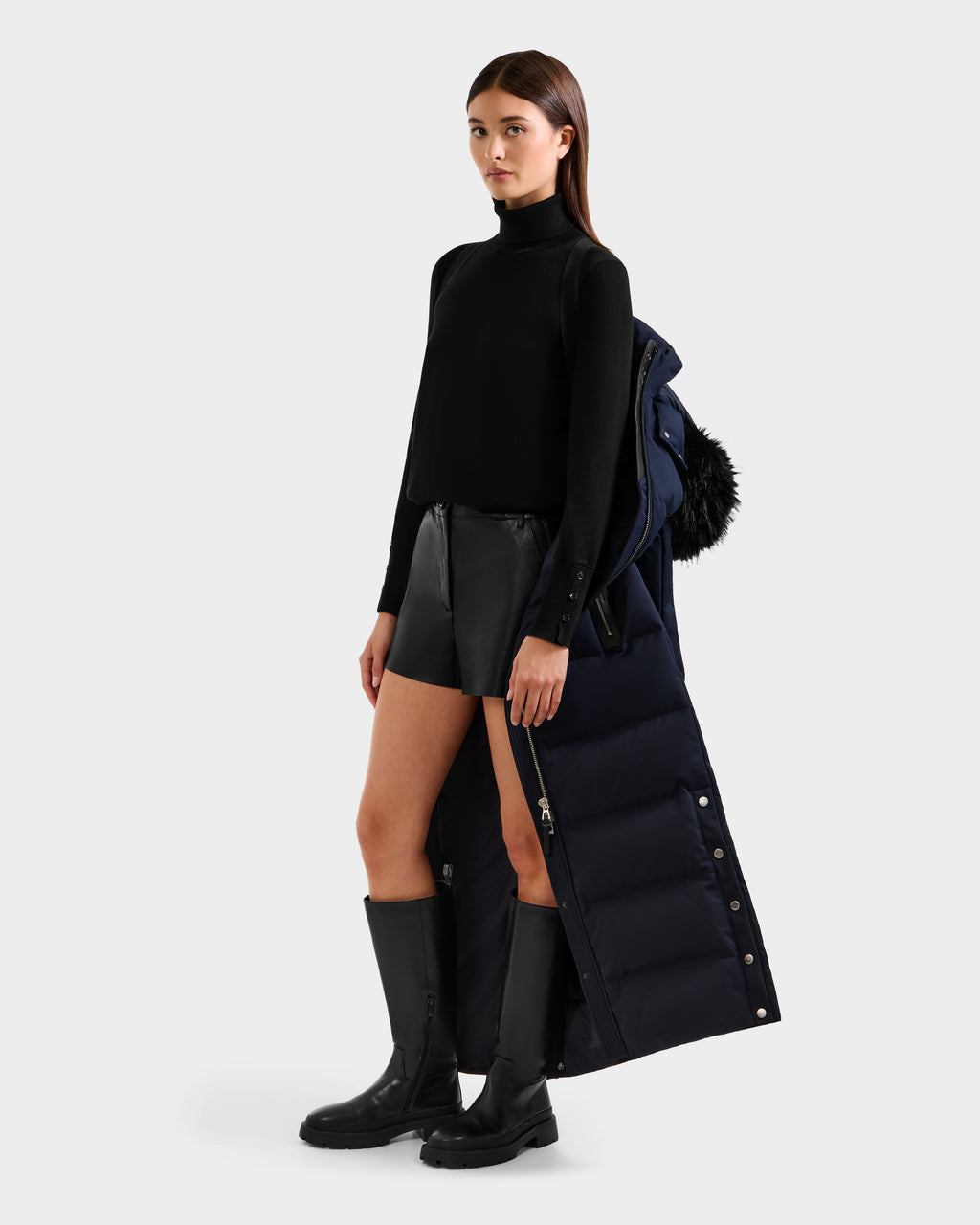 A woman in a black turtleneck, leather shorts, and knee-high boots stands holding the Rudsak IZABELLA H - 8125644 Navy puffer draped over her arm against a plain light gray background.