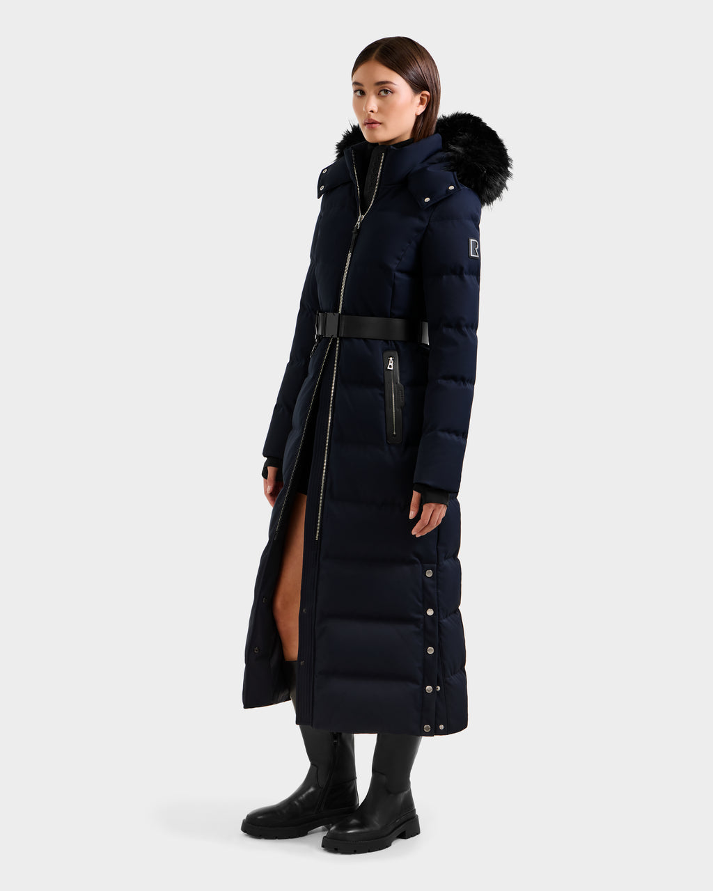 A woman stands at a slight angle against a plain light background, wearing the Rudsak IZABELLA H - 8125644 Navy puffer with a belted waist, faux fur-trimmed hood, and black boots.