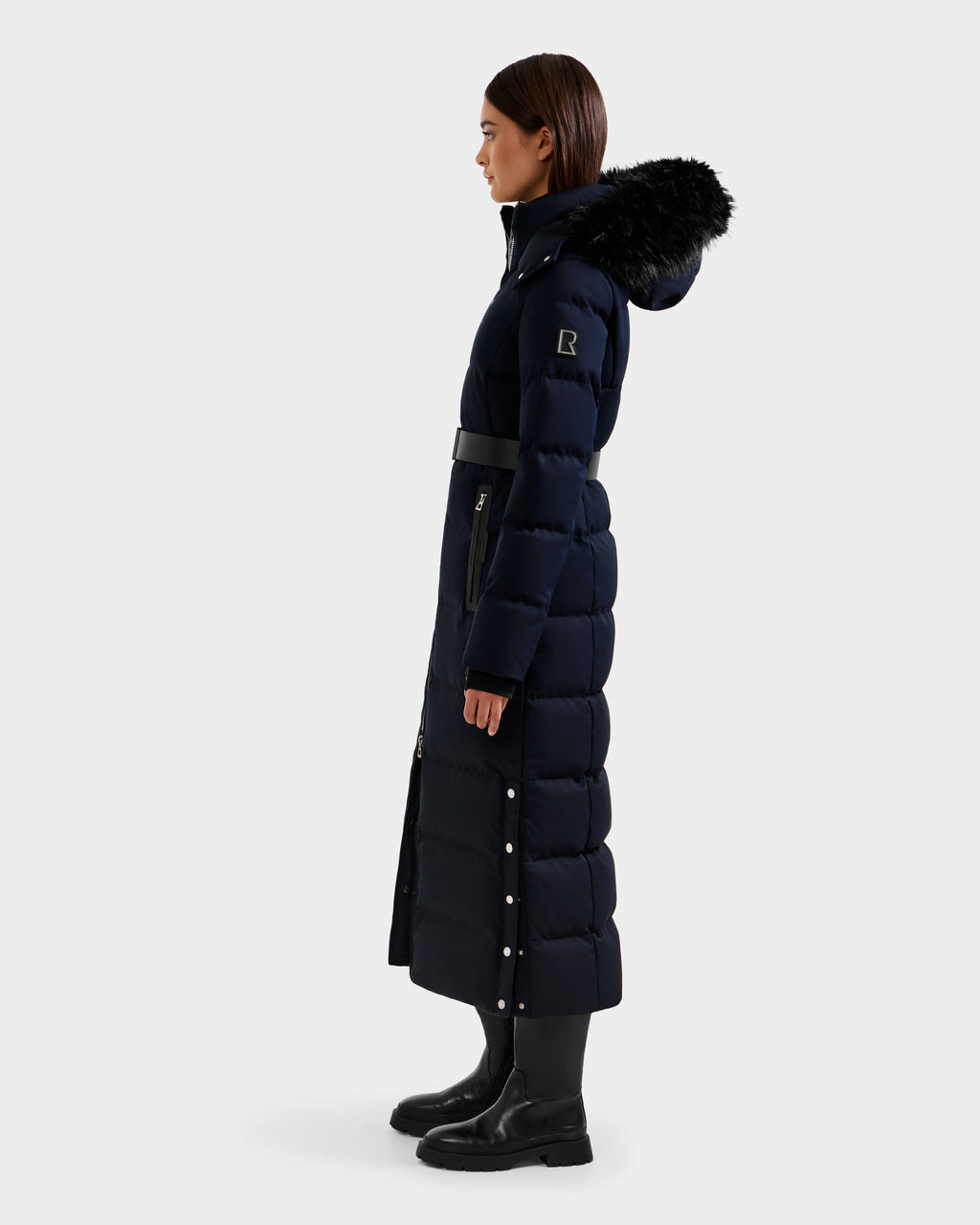 A woman stands in profile against a plain light background, wearing the Rudsak IZABELLA H - 8125644 Navy women's long puffer down coat with belted waist, fur-trimmed hood, side snap buttons, zipper pockets, and black boots.