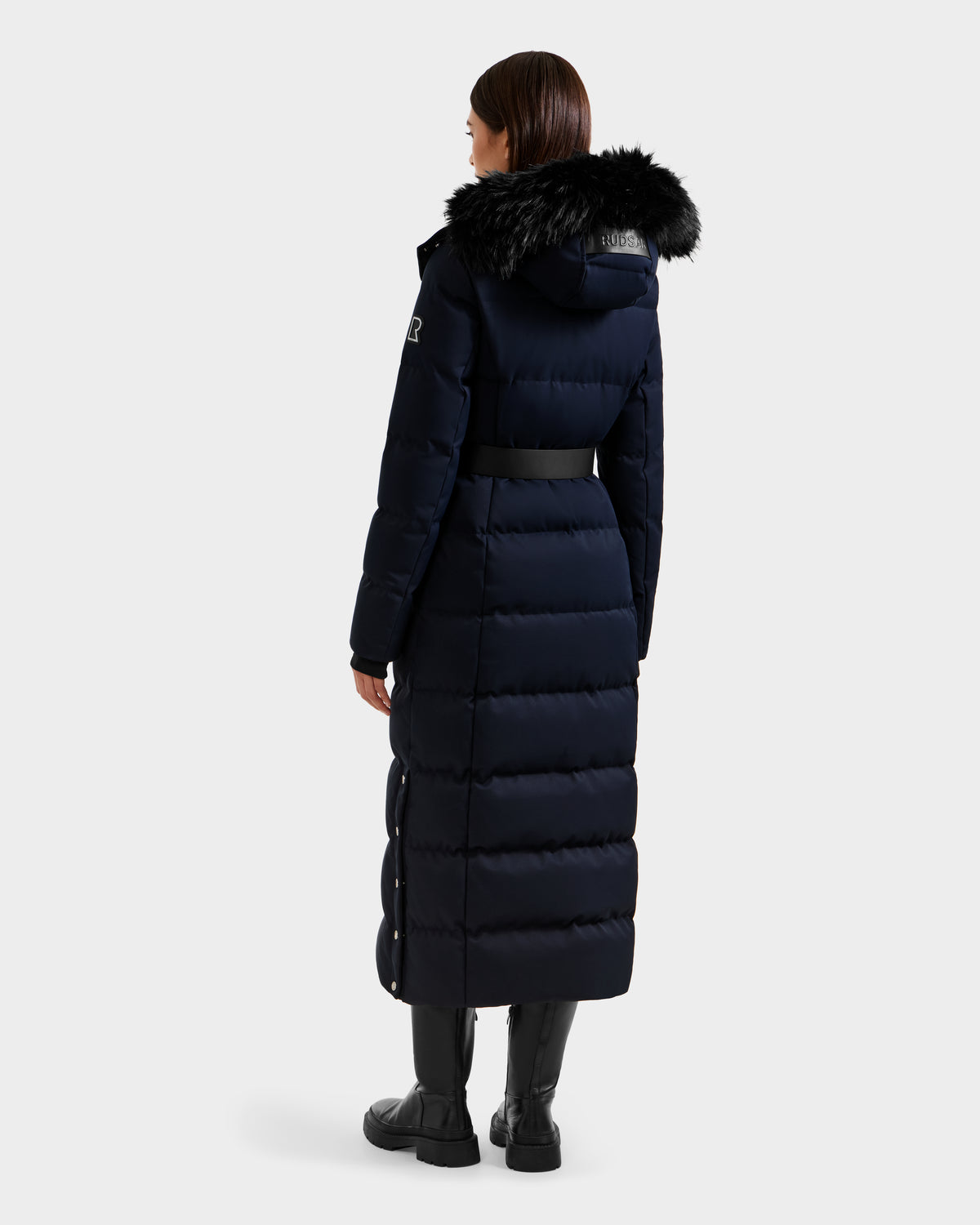A woman faces away, wearing the Rudsak IZABELLA H - 8125644 Navy down coat with a large black faux-fur hood trim, black belt, and boots. This women's puffer features horizontal stitching and a mid-calf length for winter warmth.
