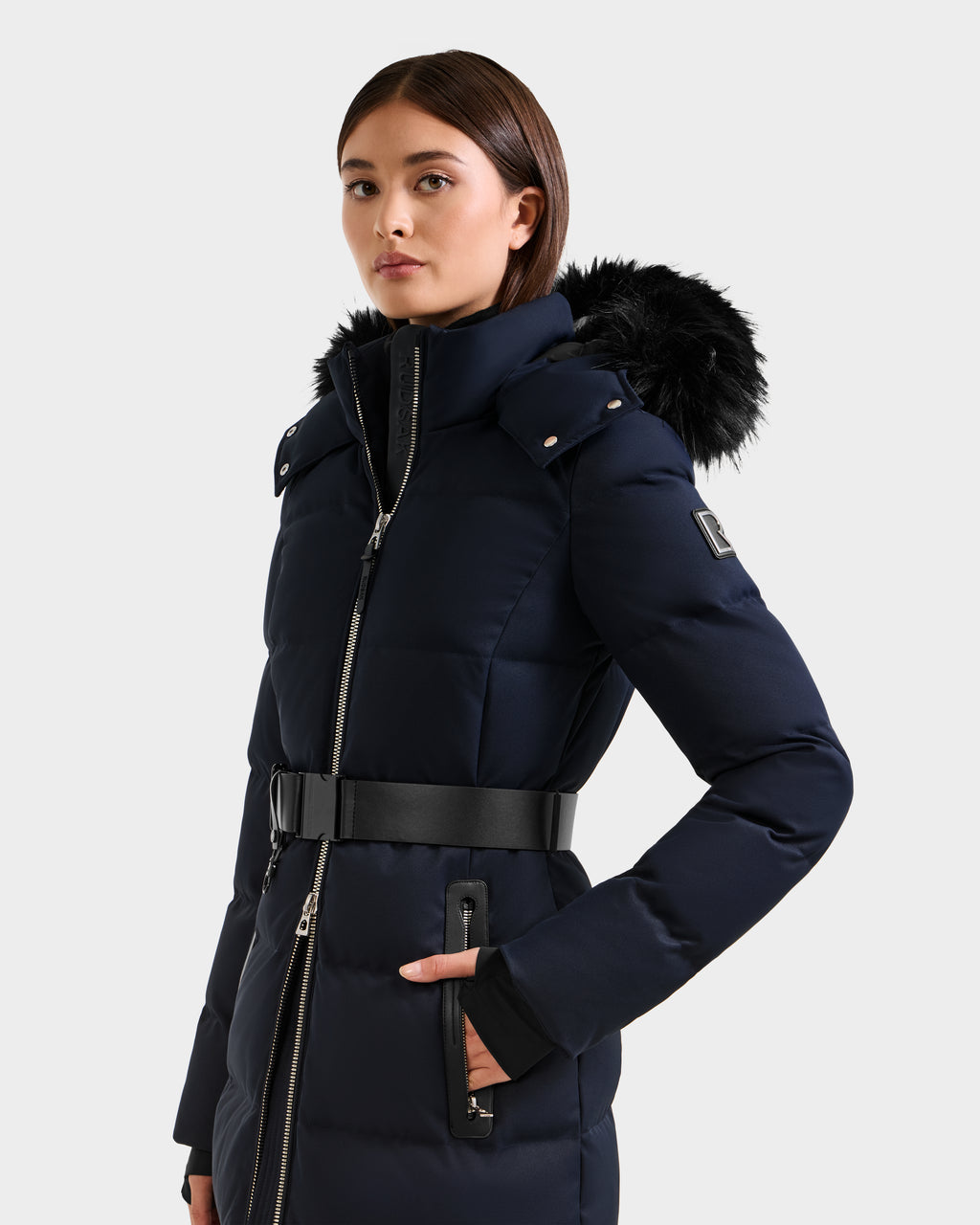 A woman wears the Rudsak IZABELLA H - 8125644 Navy belted cold-weather coat with a fur-trimmed hood, standing with one hand in her pocket against a plain light background.