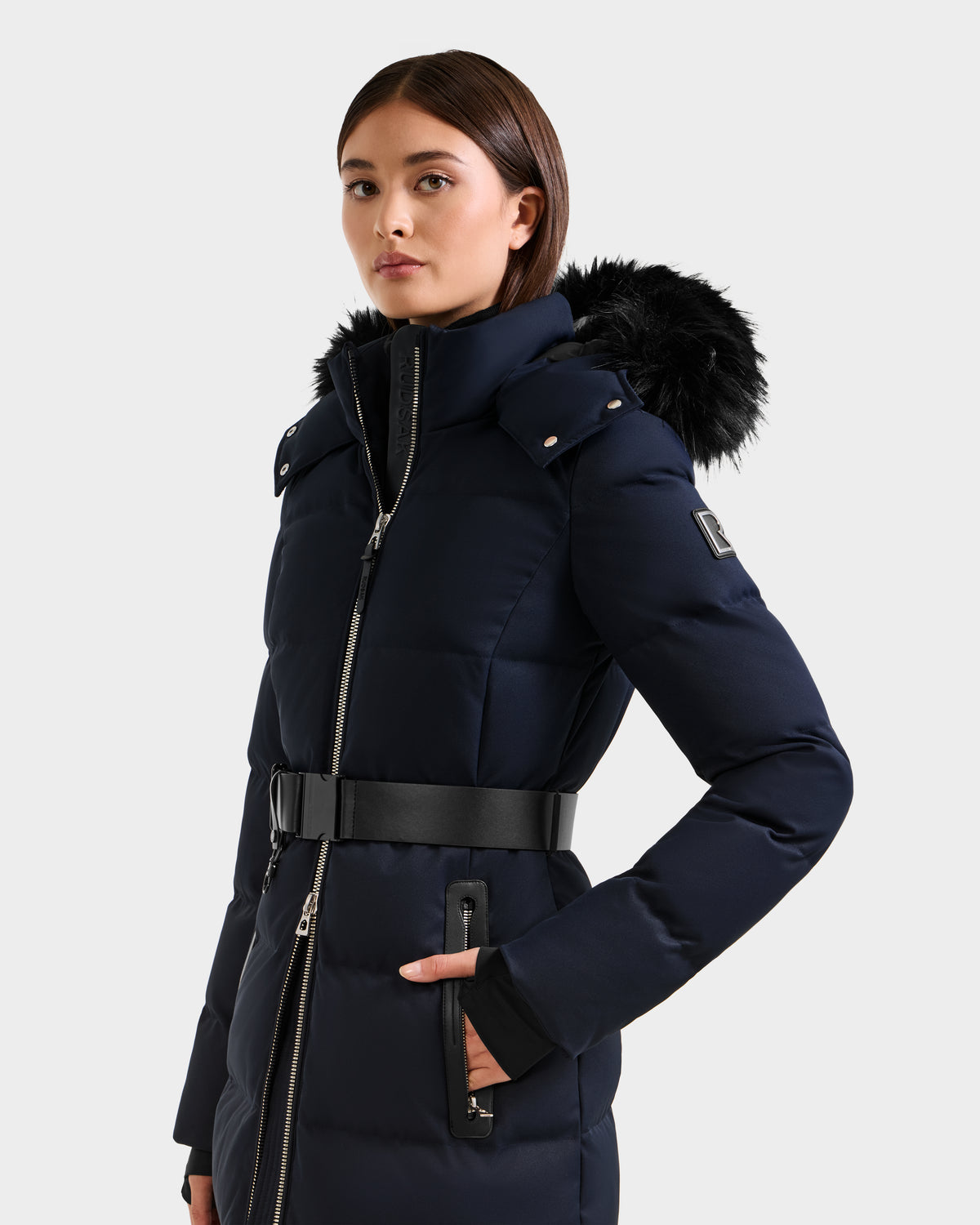 A woman wears the Rudsak IZABELLA H - 8125644 Navy belted cold-weather coat with a fur-trimmed hood, standing with one hand in her pocket against a plain light background.