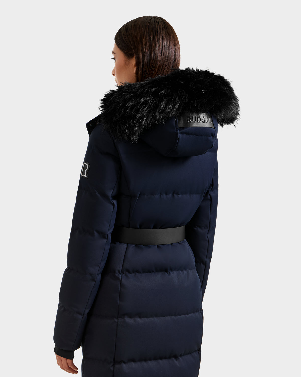 A woman with straight brown hair displays the back of the Rudsak IZABELLA H - 8125644 Navy puffer jacket, highlighting its belted waist, faux fur-trimmed hood with lettering, and an arm patch.