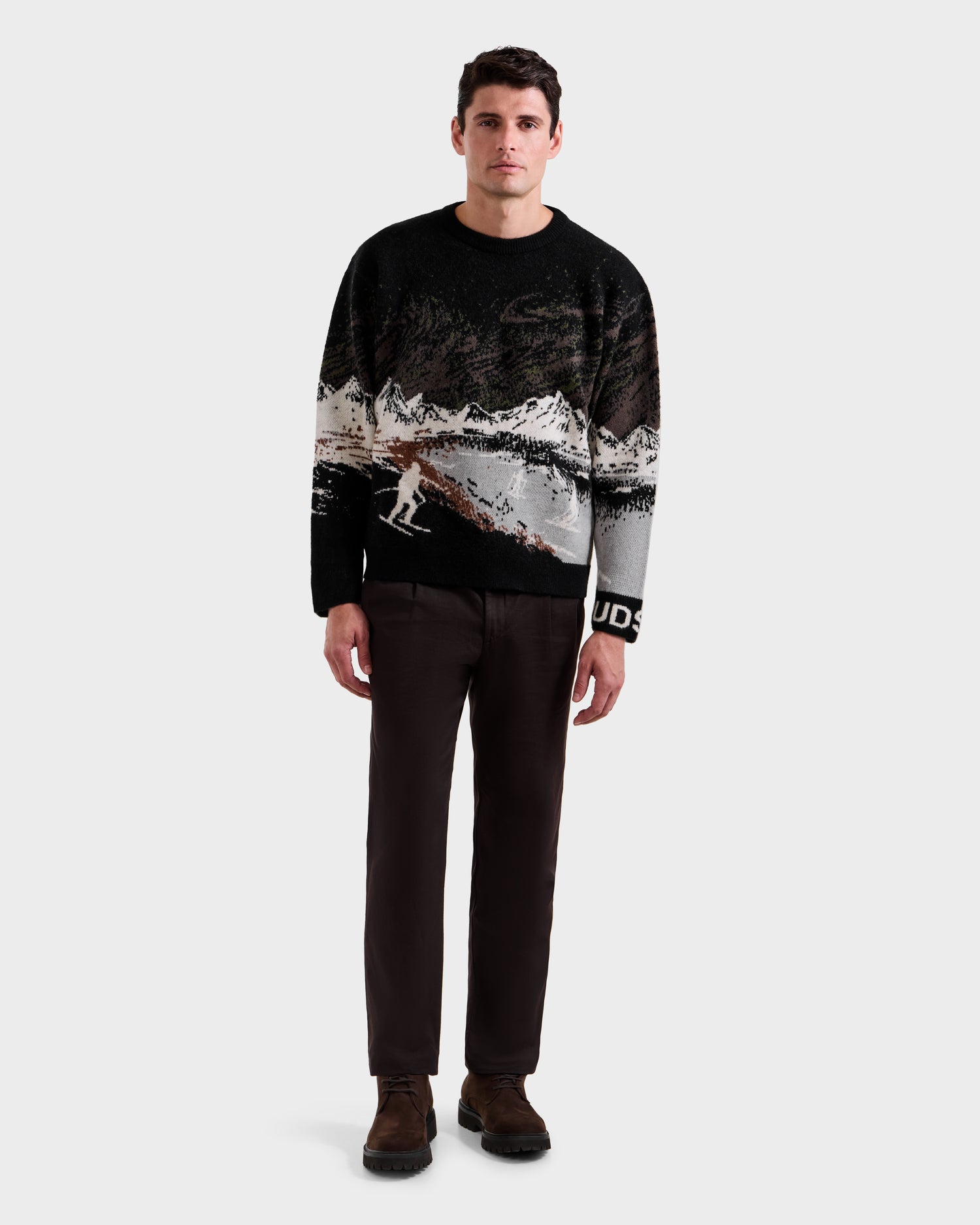 A man faces forward wearing the Rudsak IZZY - 9525557 Black M unisex knit sweater with a black and white ski mountain motif, paired with dark brown pants and brown shoes against a plain light grey background.