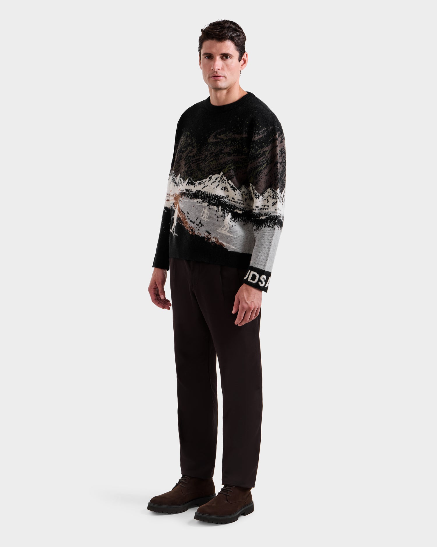 A man faces forward against a plain background wearing the Rudsak IZZY - 9525557 Black M unisex knit sweater with a ski mountain motif, dark pants, and brown shoes, channeling effortless après-ski chic.