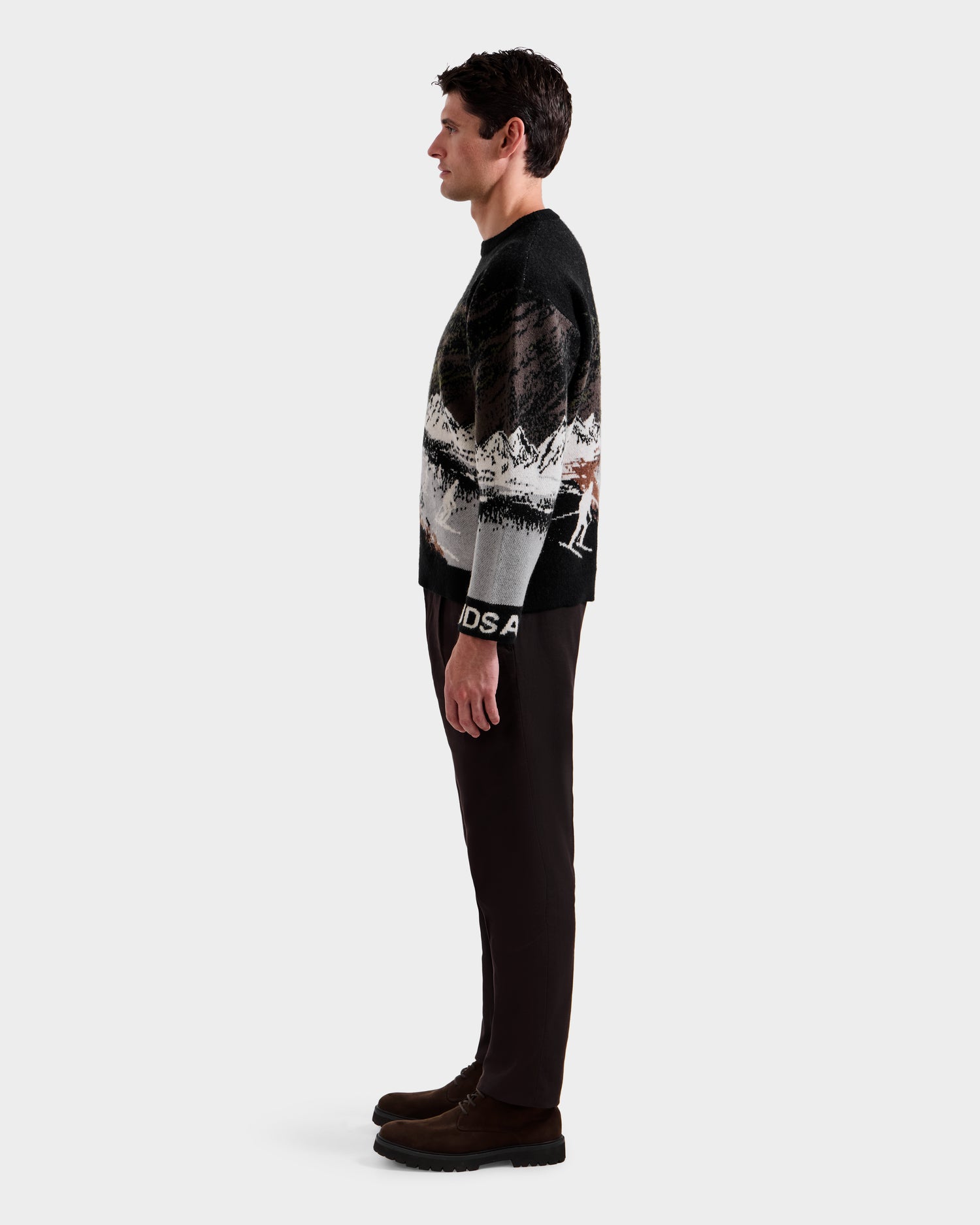 A man in profile against a plain background wears the Rudsak IZZY - 9525557 Black M unisex knit sweater with a ski mountain motif, paired with dark pants and dark brown shoes.