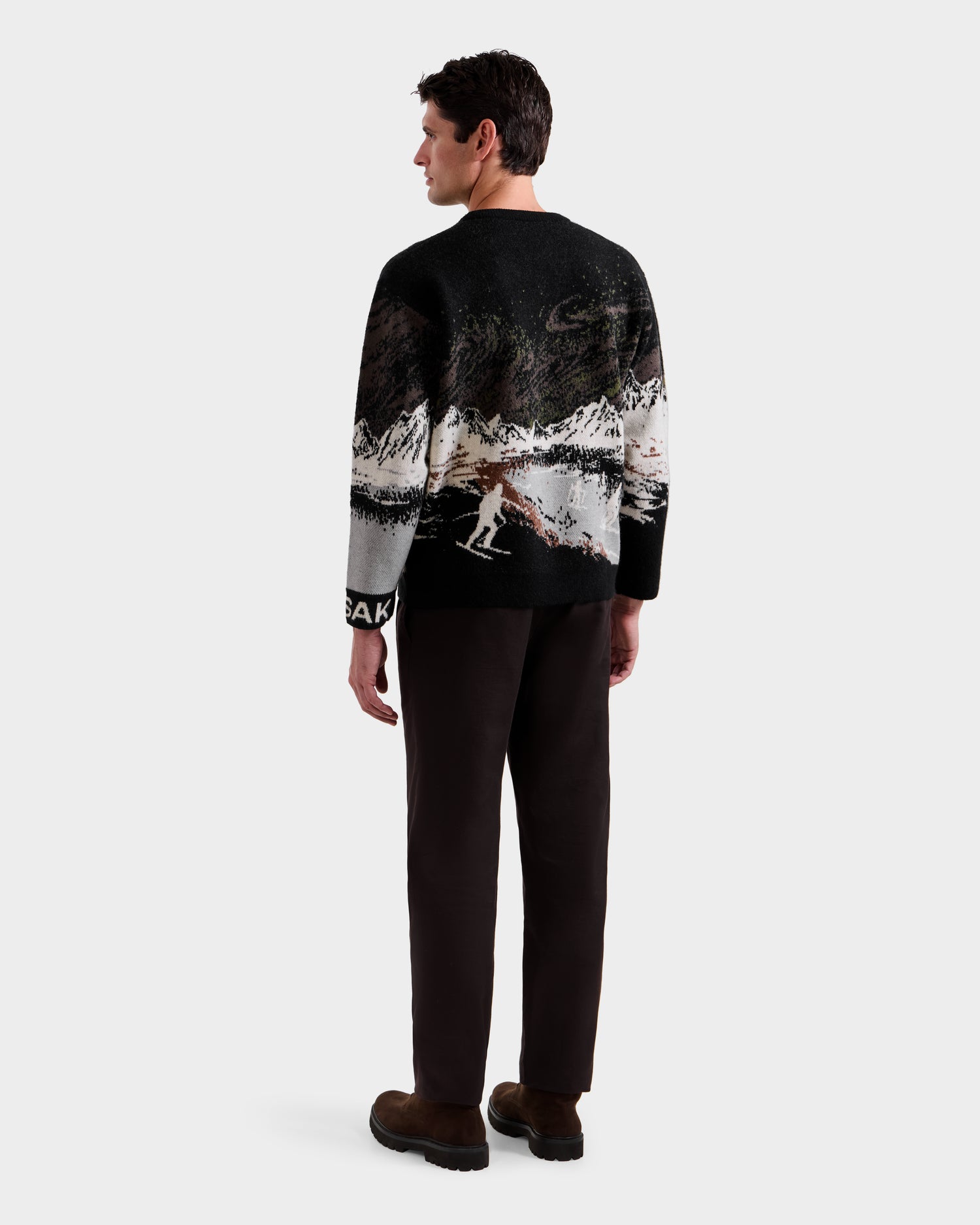 A man stands facing away, wearing the Rudsak IZZY - 9525557 Black M unisex knit sweater with a ski mountain motif, dark pants, and brown shoes against a plain light background.