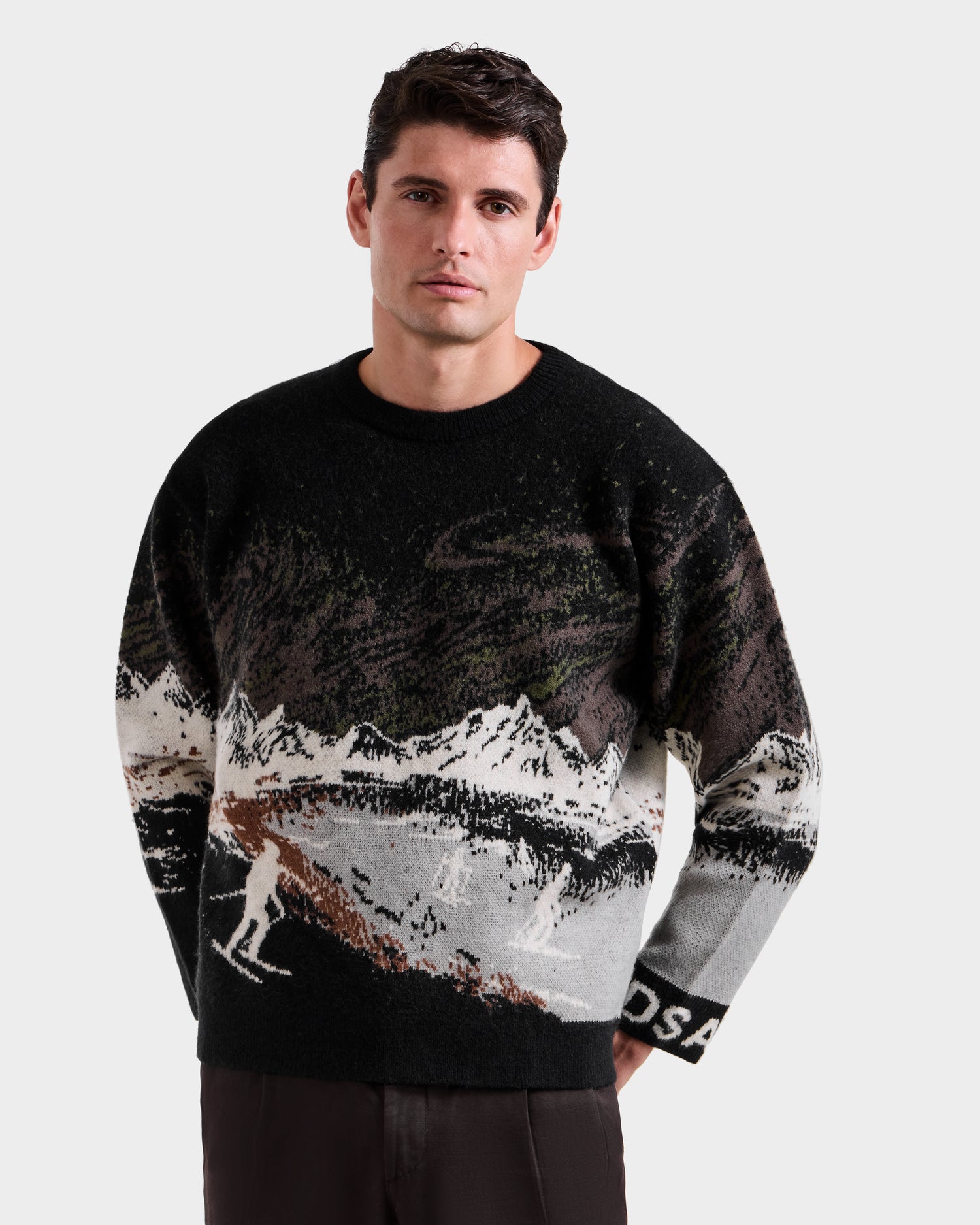 A man models the Rudsak IZZY - 9525557 Black M unisex knit sweater with a winter mountain and skier design, capturing après-ski style against a white background.