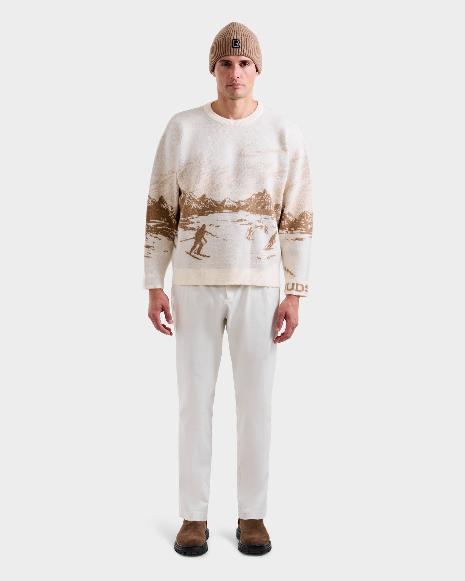 A man faces forward in a Rudsak IZZY - 9525557 Iceland M après-ski sweater with brown mountain and skier patterns, paired with a beige knit beanie, white pants, and brown shoes against a plain white background.
