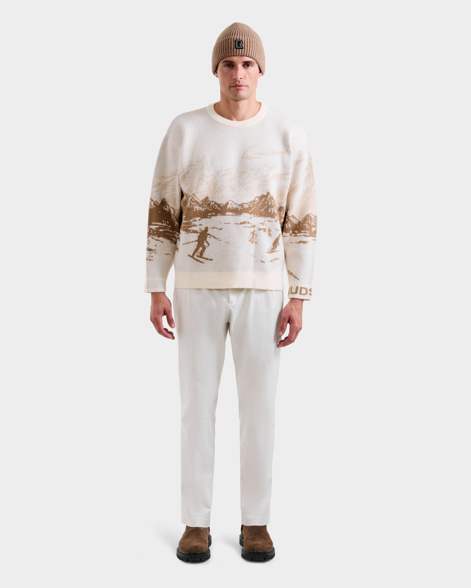 A man faces forward in a Rudsak IZZY - 9525557 Iceland M après-ski sweater with brown mountain and skier patterns, paired with a beige knit beanie, white pants, and brown shoes against a plain white background.