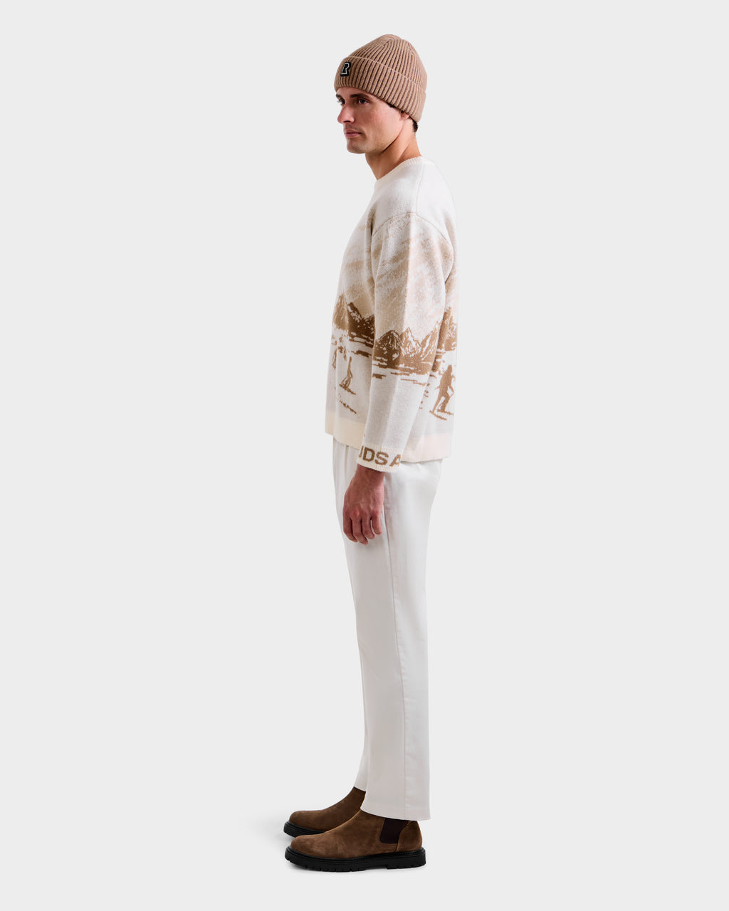 A man in profile wears the Rudsak IZZY - 9525557 Iceland M crewneck sweater with a scenic pattern, a beige beanie, off-white pants, and brown suede boots against a plain light background.