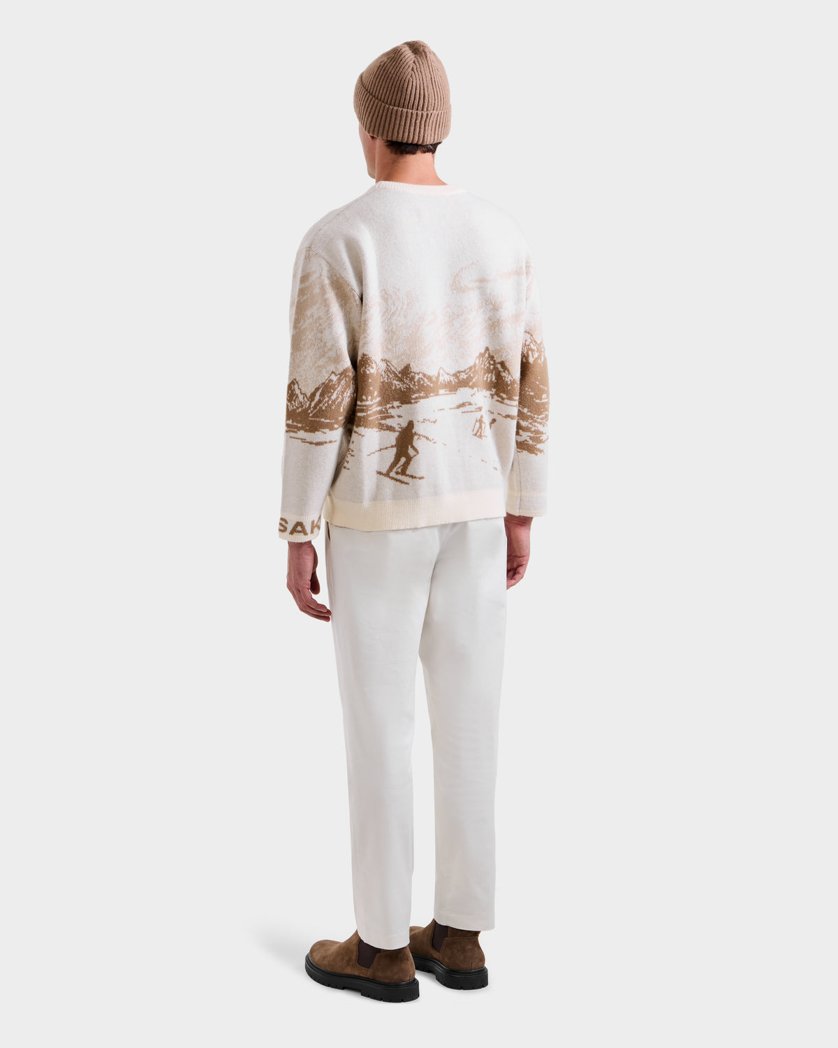 A person wearing the Rudsak IZZY - 9525557 Iceland M beige beanie, a white après-ski sweater with a brown landscape and skier design, white pants, and brown shoes stands with their back to the camera against a plain white background.