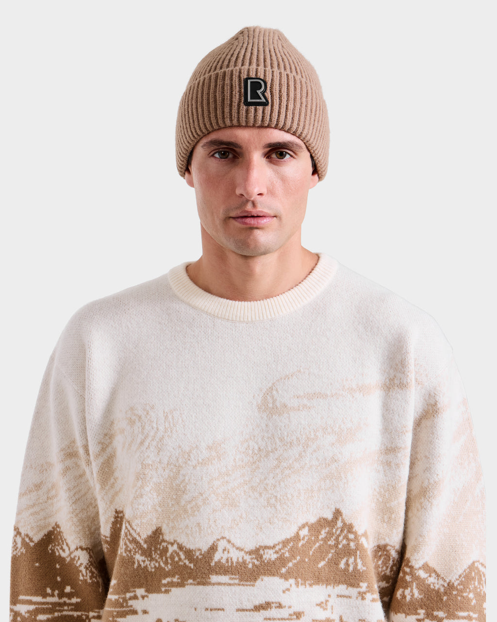 A man models the Rudsak IZZY - 9525557 Iceland M beige knit beanie with a black B logo and a cream sweater featuring a brown mountain design, capturing après-ski style against a simple light backdrop.