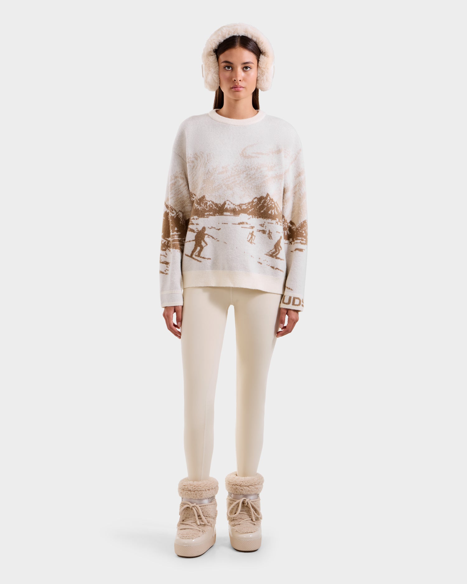 A woman stands facing forward against a white background, wearing Rudsak's IZZY - 9525557 Iceland W: a cream and brown unisex ski-motif knit sweater, paired with fluffy earmuffs, cream leggings, and beige fur-lined winter boots.