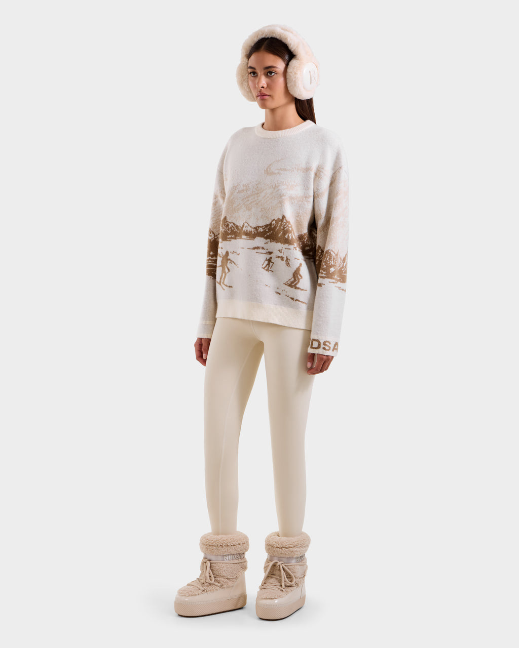 A person wearing Rudsak’s IZZY - 9525557 Iceland W boots, cream earmuffs, a unisex knit sweater with a brown ski mountain motif, and cream leggings stands against a plain light background.