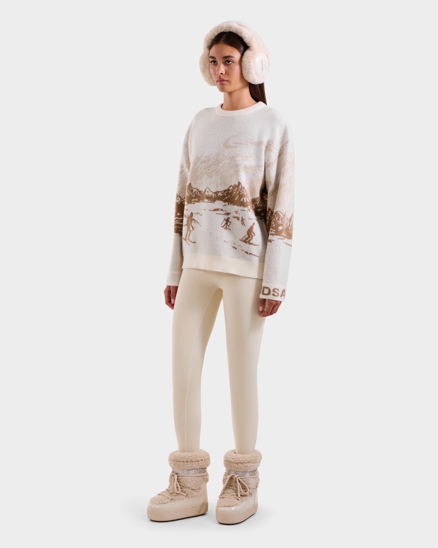 A person wearing Rudsak’s IZZY - 9525557 Iceland W boots, cream earmuffs, a unisex knit sweater with a brown ski mountain motif, and cream leggings stands against a plain light background.