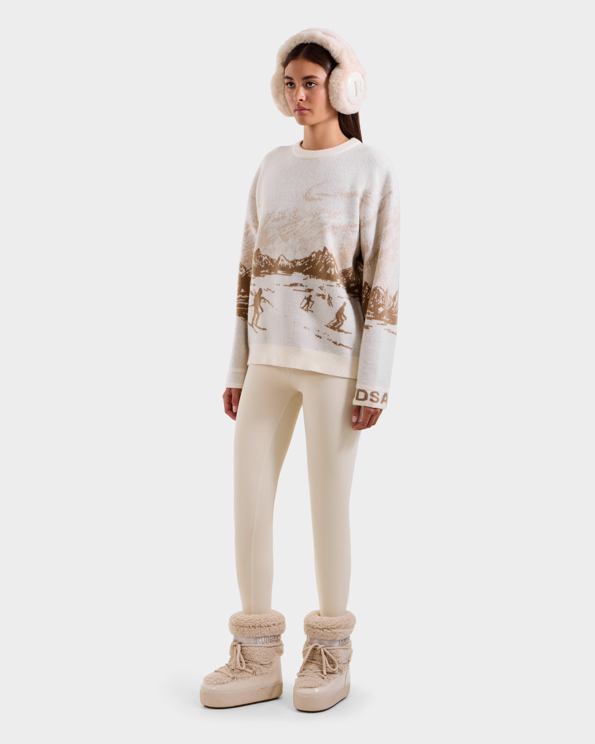 A person wearing Rudsak’s IZZY - 9525557 Iceland W boots, cream earmuffs, a unisex knit sweater with a brown ski mountain motif, and cream leggings stands against a plain light background.