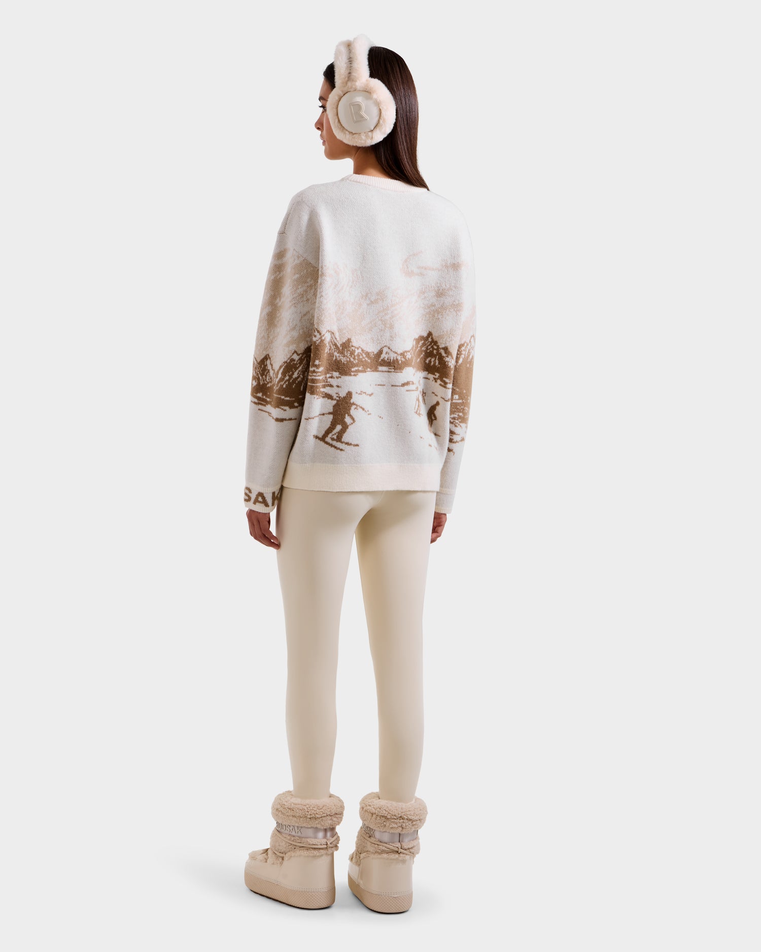 A woman faces away, wearing Rudsak’s IZZY - 9525557 Iceland W: a cozy unisex knit sweater with a ski mountain motif, paired with cream earmuffs, leggings, and beige fur-lined boots, set against a plain white background.