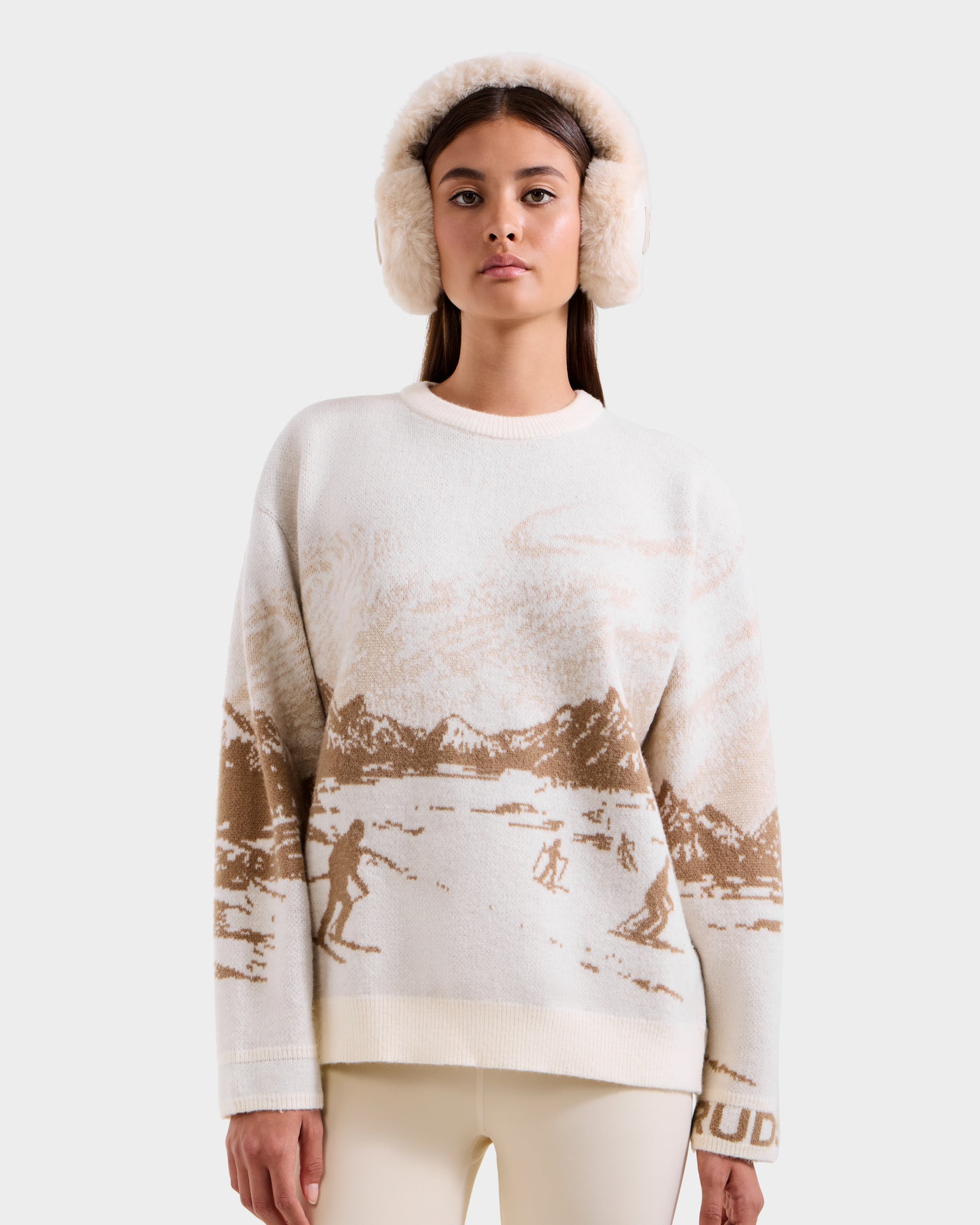A woman in Rudsak’s IZZY - 9525557 Iceland W cream unisex knit sweater with a brown ski mountain motif and fluffy earmuffs stands against a light background, exuding après-ski chic.