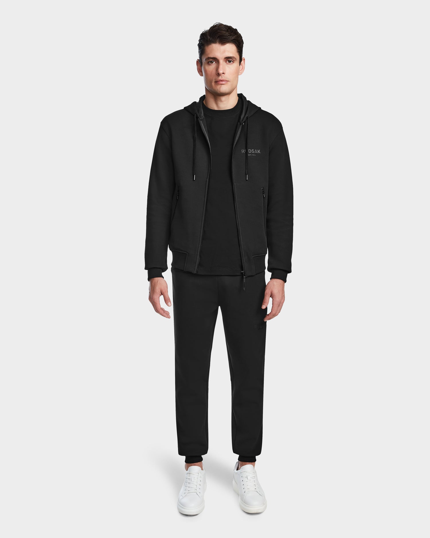 A man stands facing forward wearing a black Rudsak JACE - 9524010 hoodie (size M), black t-shirt, black jogger pants, and white sneakers against a plain white background.