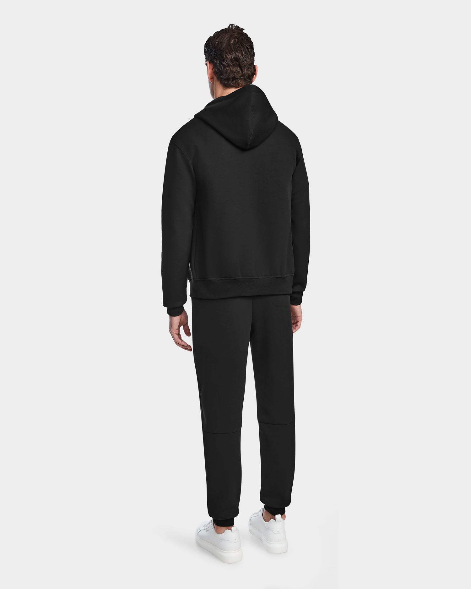 A man faces away from the camera, wearing the Rudsak JACE - 9524010 Black M hoodie and matching sweatpants with white sneakers, set against a plain white background.