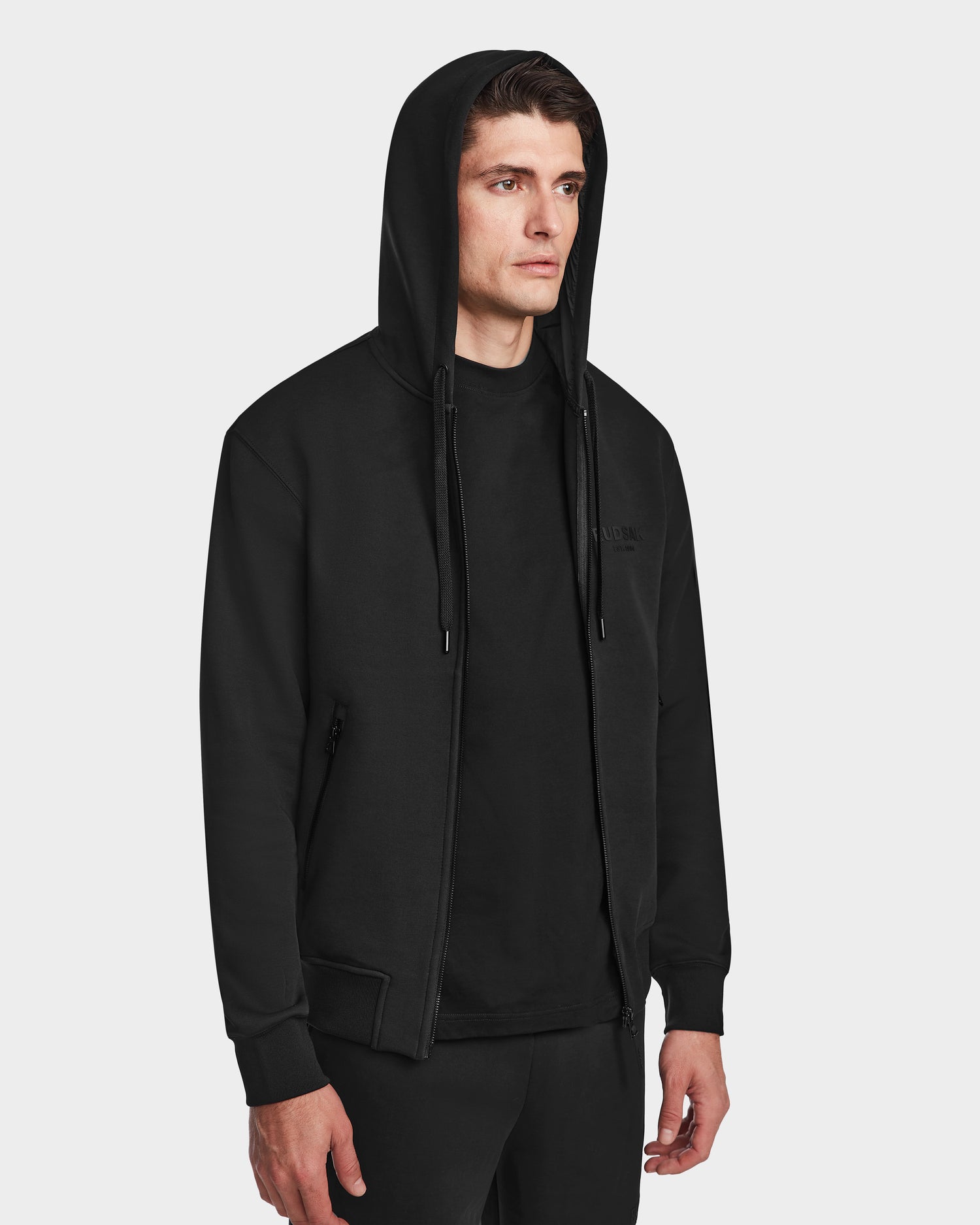 A man stands slightly turned against a plain white background, wearing the Rudsak JACE - 9524010 Black M hoodie over a black shirt.