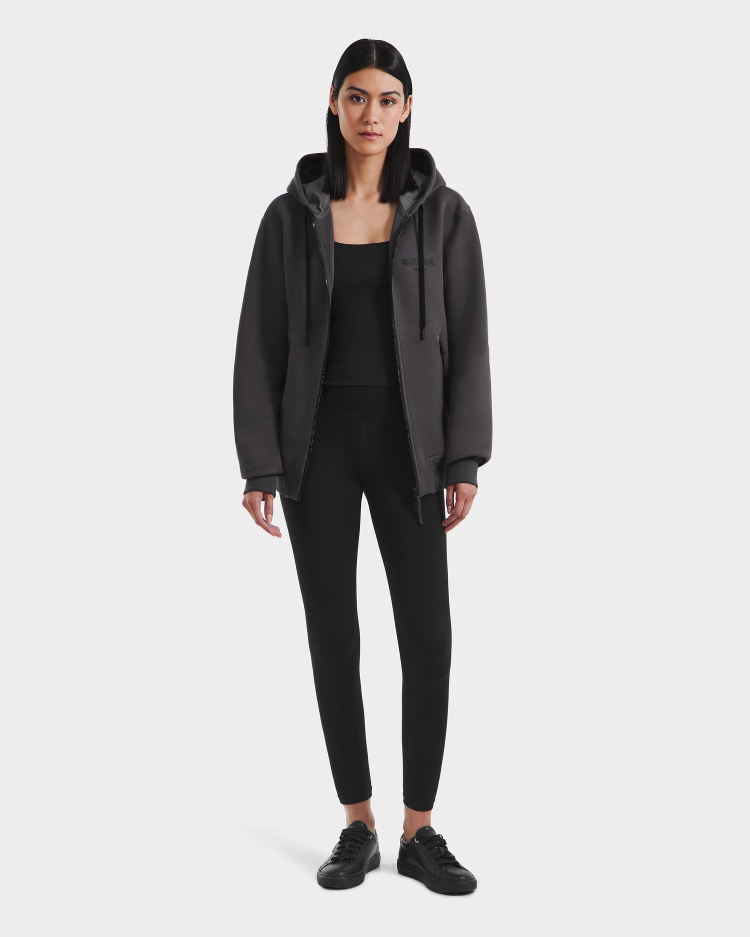 A woman with straight dark hair wears the Rudsak JACE - 9524010 Grey W hoodie over a black top, paired with black leggings and sneakers, standing against a white background.