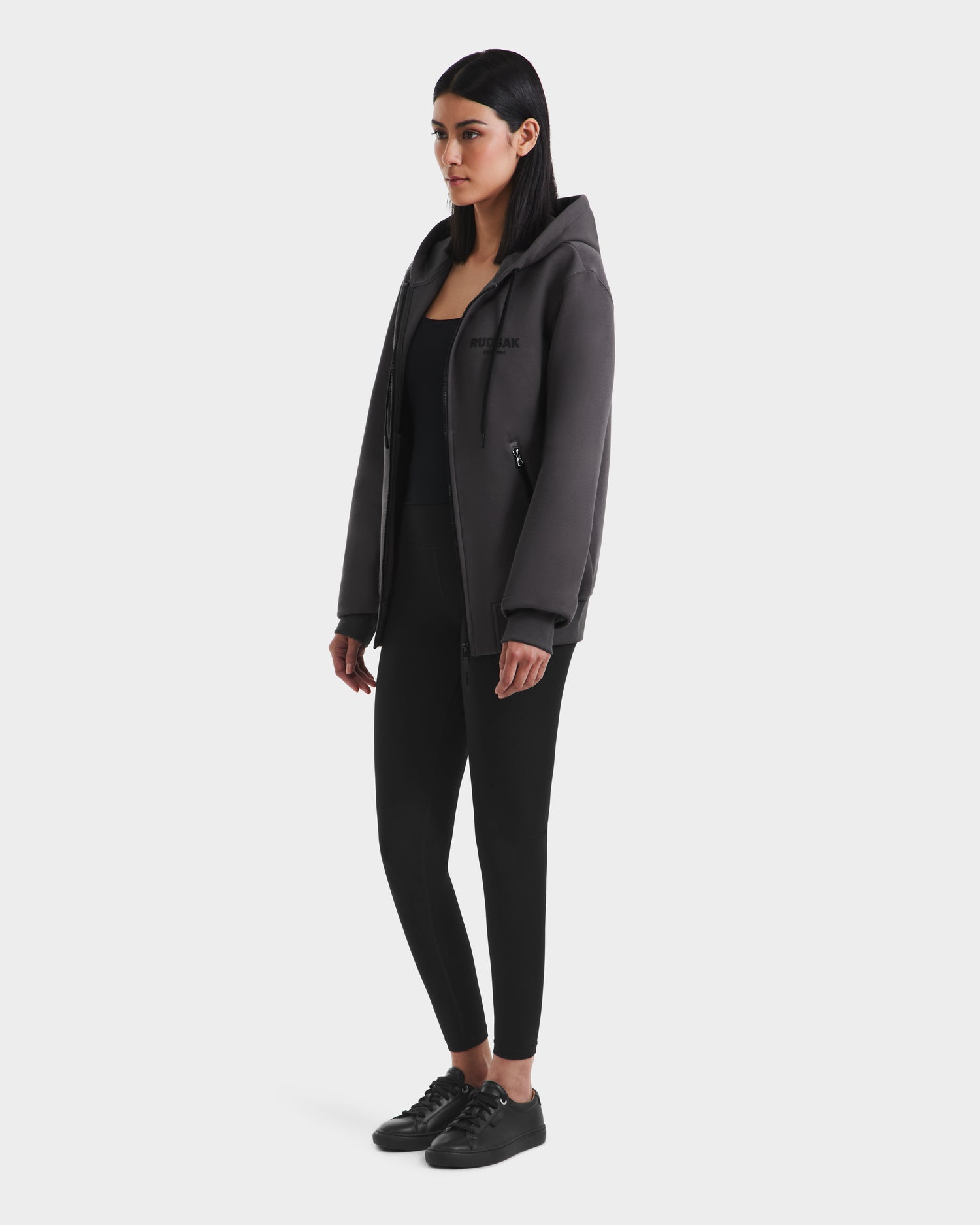 A woman with straight dark hair wears the Rudsak JACE - 9524010 Grey W hoodie, paired with black leggings and black sneakers, standing against a plain white background.