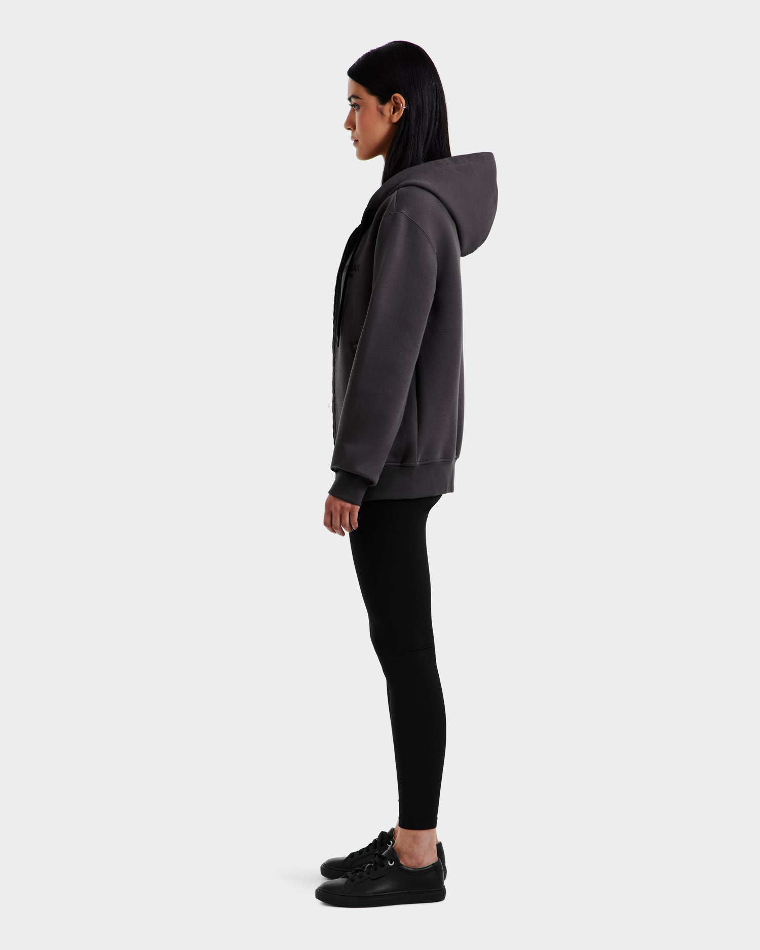 A woman in profile faces left, wearing a Rudsak JACE - 9524010 Grey W hoodie, black leggings, and black sneakers against a plain light gray background.