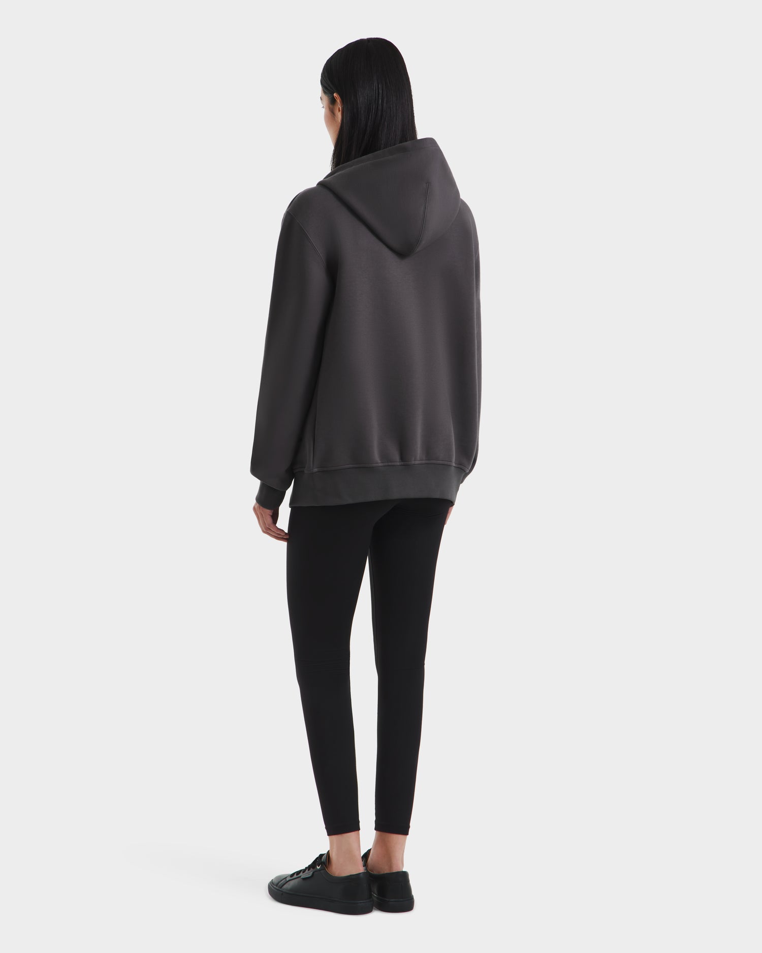 A person with long dark hair stands facing away in a Rudsak JACE - 9524010 Grey W hoodie, black leggings, and black sneakers against a plain light background.