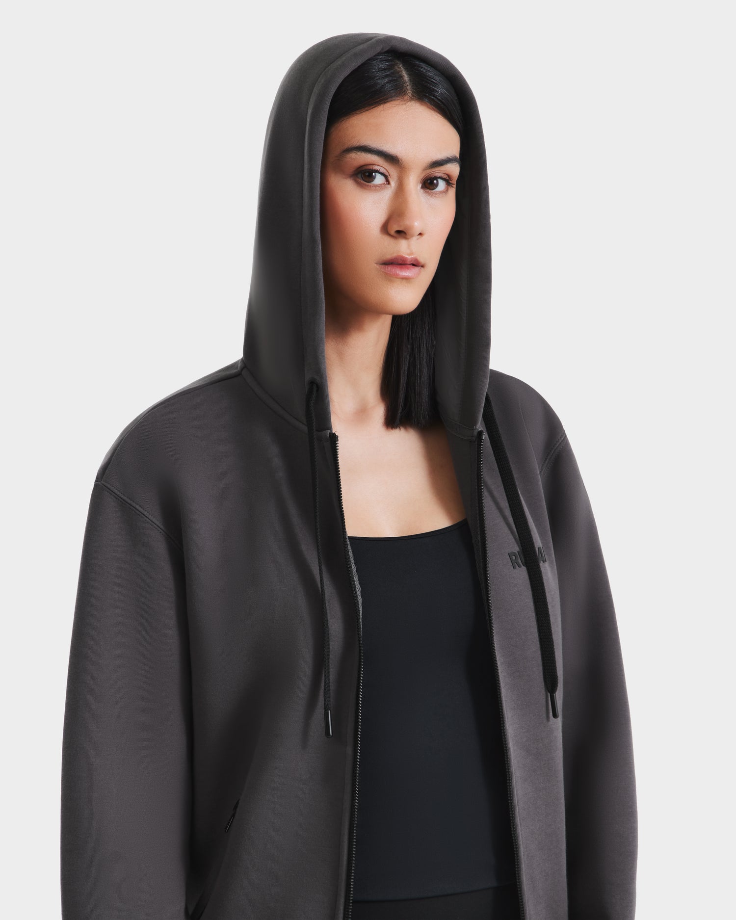 A woman with straight dark hair wears the Rudsak JACE - 9524010 Grey W hoodie, crafted from a premium cotton blend, over a black top. The hood is up as she looks to the side with a neutral expression against a plain white background.