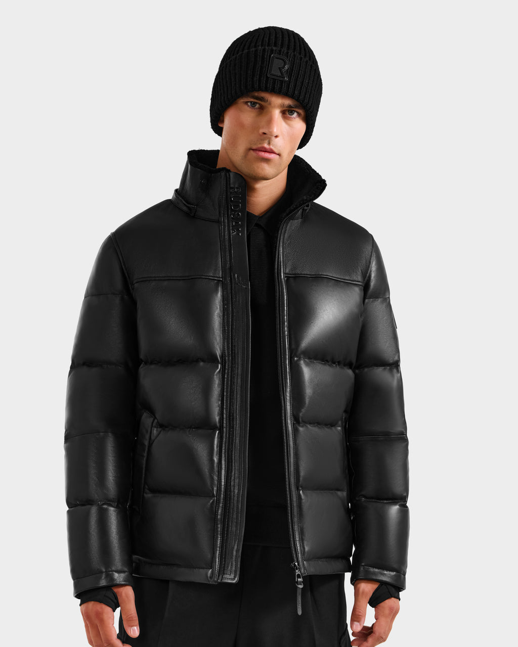 A man wearing the Rudsak JAMESON - 6125678 Black men's winter jacket with a black knit beanie and black pants stands against a plain light gray background, looking directly at the camera.