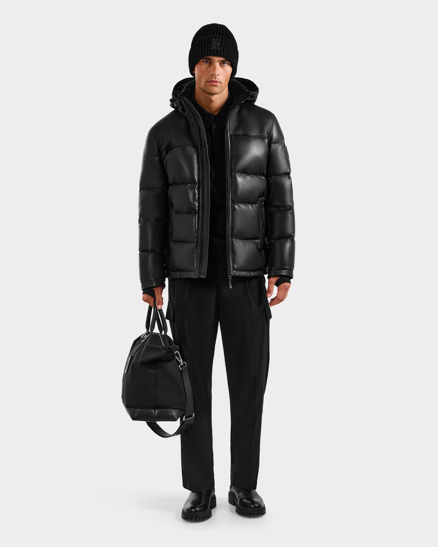 A person stands against a white background, holding the Rudsak JAMESON - 6125678 Black duffel bag and wearing a black beanie, quilted leather down jacket, black hoodie, black pants, and black shoes.