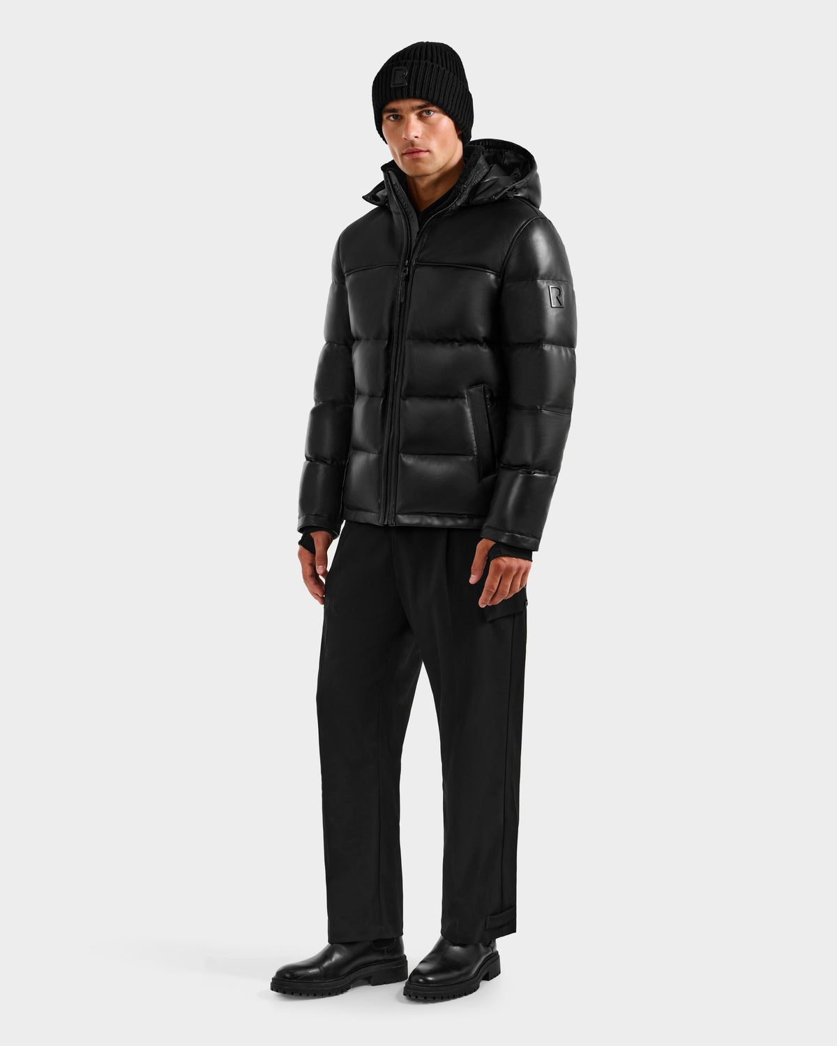 Wearing the Rudsak JAMESON - 6125678 Black men's leather down jacket, a person pairs it with a black beanie, black pants, and black shoes, standing against a light gray background for a classic winter look.