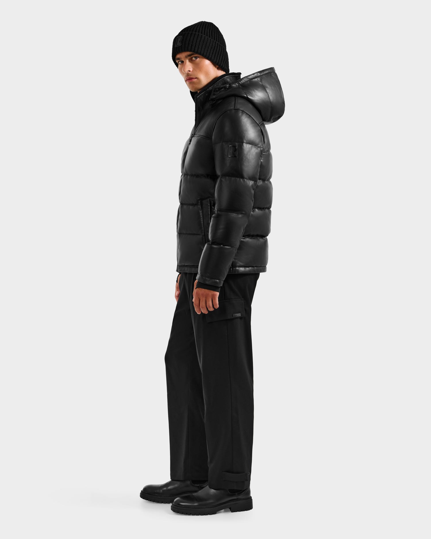 A man stands in profile wearing the Rudsak JAMESON - 6125678 Black leather down jacket with hood, styled with a black beanie, pants, and boots against a plain light background—a true winter essential.