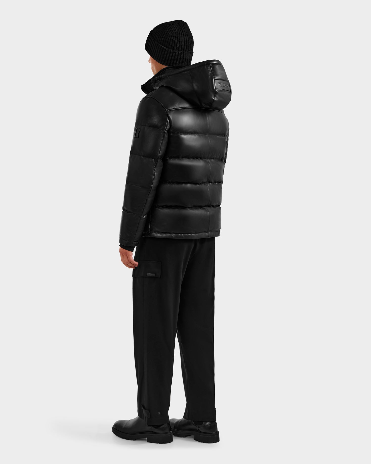 A person faces away, wearing the Rudsak JAMESON - 6125678 Black puffer jacket with a hood, black pants, boots, and a black knit beanie—classic winter essentials against a plain light gray background.