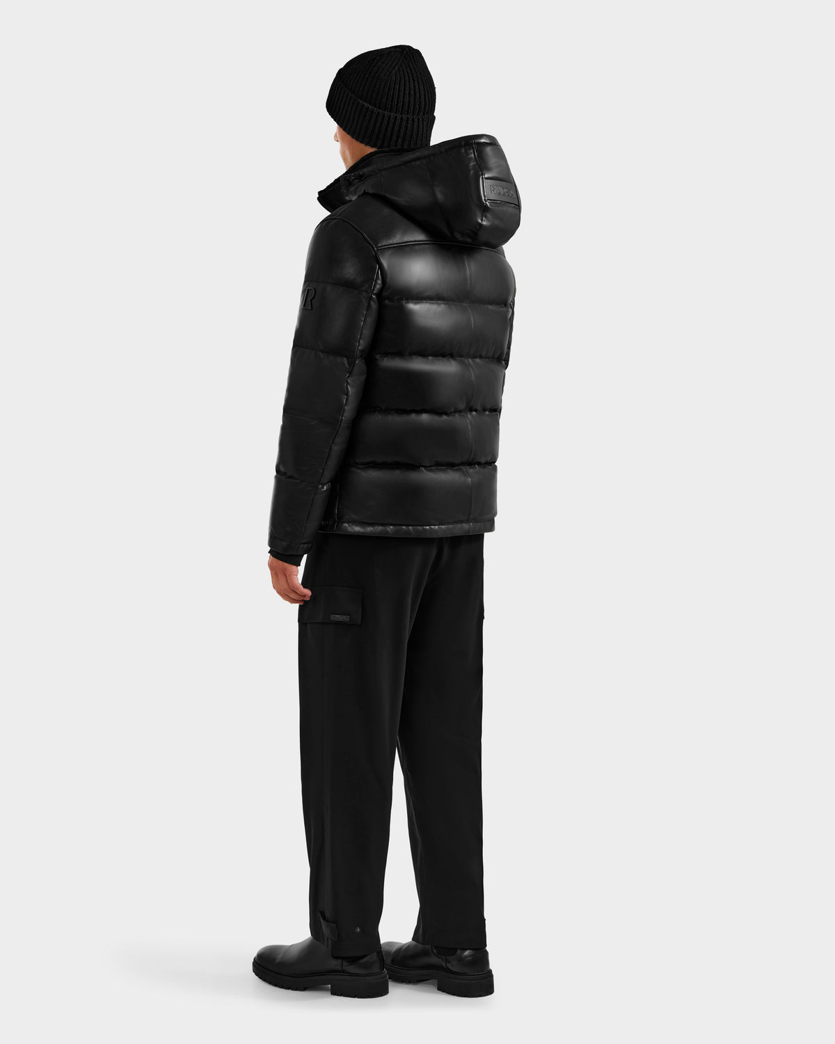 A person faces away, wearing the Rudsak JAMESON - 6125678 Black puffer jacket with a hood, black pants, boots, and a black knit beanie—classic winter essentials against a plain light gray background.