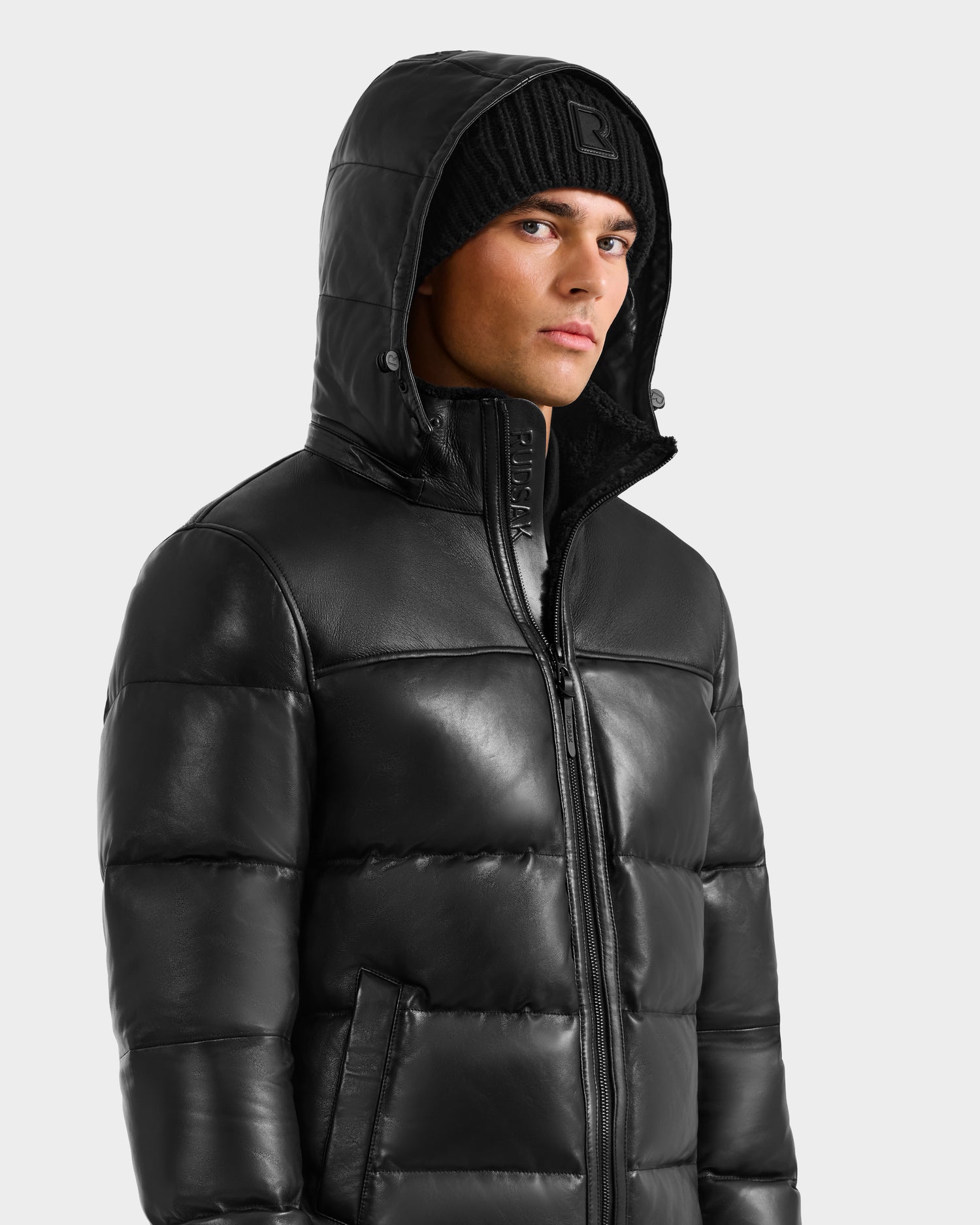 A man stands slightly to the side against a plain light background, wearing the Rudsak JAMESON - 6125678 Black quilted leather down jacket with high collar and hood, paired with a black knit beanie.