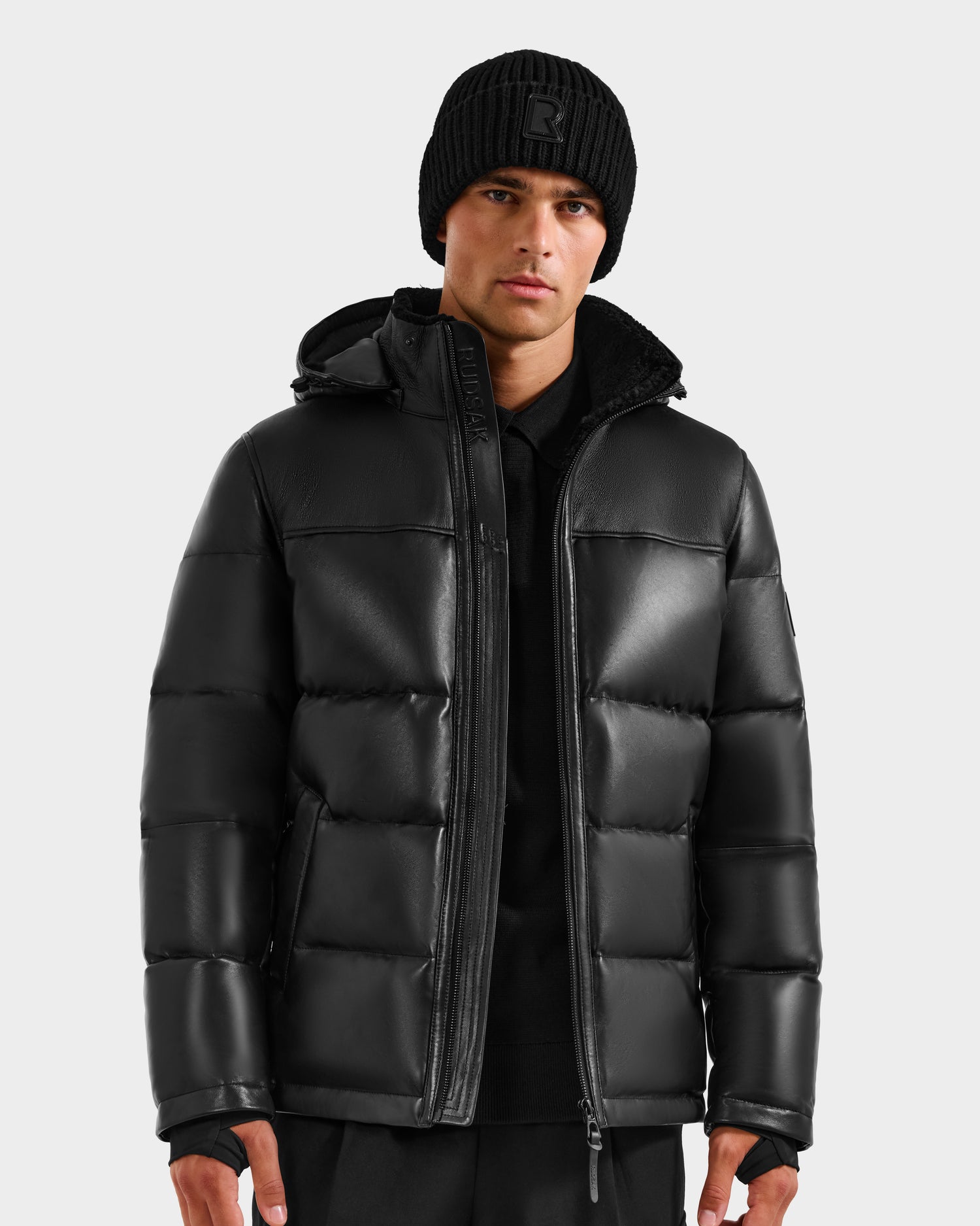 A person wears the Rudsak JAMESON - 6125678 Black men’s quilted leather down jacket with a hood, paired with a black knit beanie and pants, standing against a light gray background—a true winter essential.