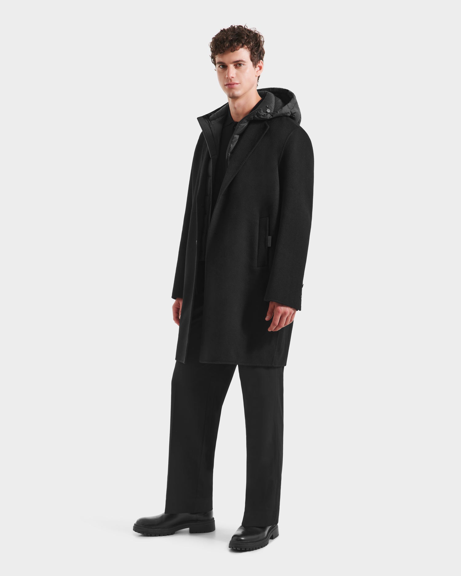 A man with short curly hair models the Rudsak JEREMIAH - 6125509 Black men’s wool-blend coat over a dark hoodie, black pants, and black shoes, standing against a plain white background.