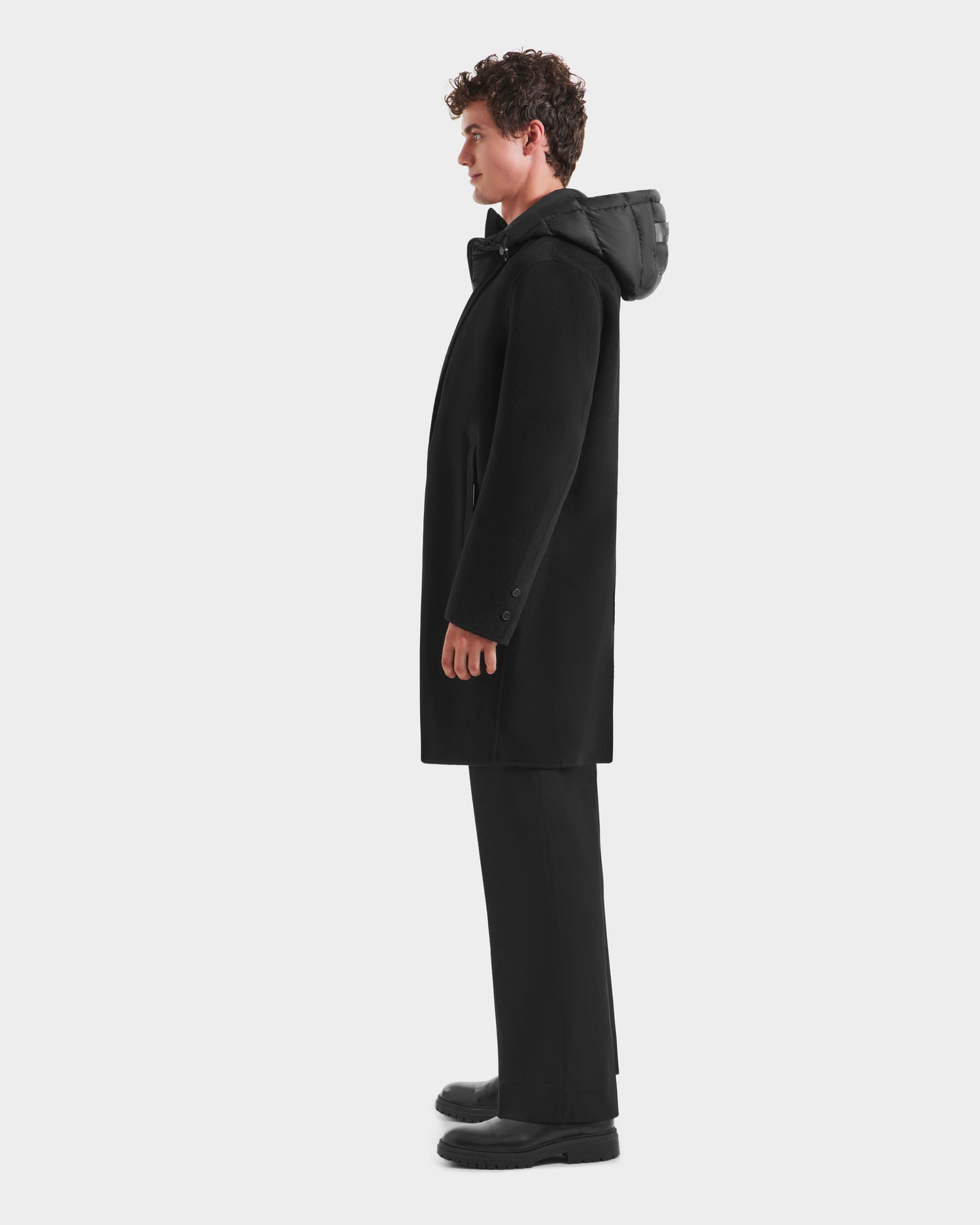 A man with curly hair stands in profile wearing the Rudsak JEREMIAH - 6125509 Black men’s wool-blend hooded coat, paired with black pants and shoes, against a plain white background—a staple look for cold weather.
